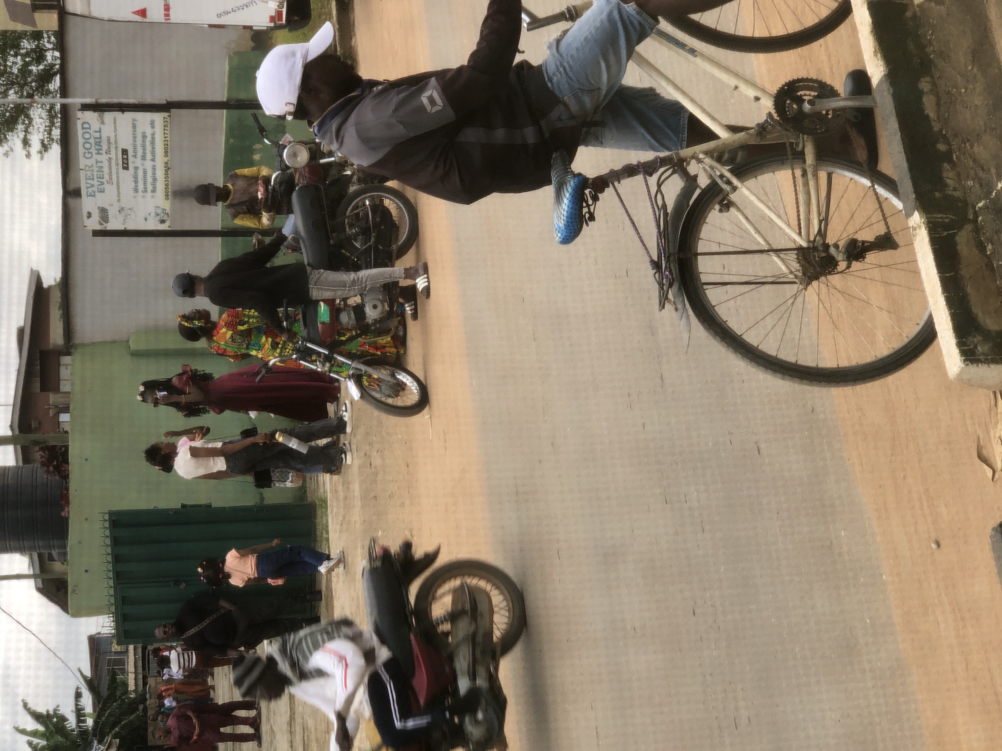 The image depicts a street scene in Lagos, Nigeria. In the foreground, a man wearing a white cap and a gray and dark jacket over denim shorts sits on a light-colored bicycle. He is on a dirt road, with a concrete barrier in the lower right corner. Behind him, other individuals and vehicles are visible. A person on a red motorcycle is partially visible on the left. Further in the background, a group of people are gathered. Some are dressed in traditional attire, while others are in casual wear. A green fence and a building with a sign that reads "EVER GOOD EVENT HALL" are prominent in the mid-ground. The sign also lists services such as "Wedding, Anniversary, Seminar, Meetings, Religious Activities, etc." and provides phone numbers. Several motorcycles are parked or in motion. The overall environment suggests an active urban setting, possibly during daytime.