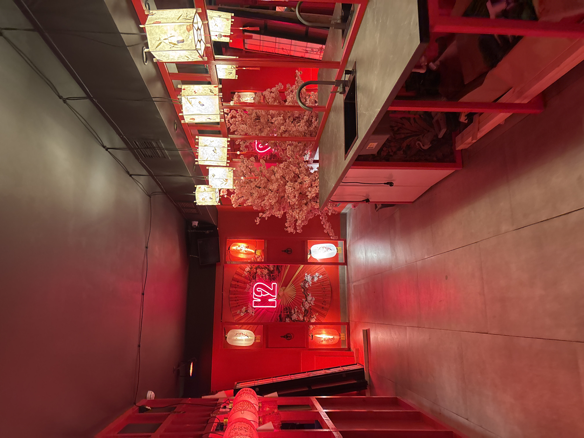 This indoor establishment, located in Saint Petersburg, Russia, presents a thematic environment dominated by a red color scheme and East Asian-inspired decor. The floor consists of light-colored, large square tiles, reflecting a red hue from the ambient lighting. The ceiling is dark grey or black, featuring exposed electrical conduit and wires, a white dome security camera in the upper left, and a black speaker box mounted in the upper right; a ventilation grate is also visible on the ceiling/upper wall on the right.
The left wall is entirely red, incorporating vertical red shelving units and several recessed sections, each containing an illuminated traditional lantern, some white and some orange/red. Further left, a cluster of hanging red traditional lanterns with white accents is visible. A dark, tall object, possibly a standing sign or partition, leans against the left wall.
The back wall is red and features a large, decorative red fan motif. Centrally placed on this fan is a prominent red neon sign displaying the letters "K2".
A long, dark grey countertop with a built-in black sink and a dark-colored single-lever faucet extends along the right side of the frame, supported by red-framed legs. Above the counter, a series of rectangular hanging lanterns with delicate artistic patterns (e.g., birds, foliage) emit a warm glow. Behind these lanterns and the counter, multiple mirrors reflect artificial pink cherry blossom trees, which are positioned on the far right. Below the counter, a white cabinet is visible on the left, and further right, unfinished storage comprising wooden planks and cardboard boxes is exposed. A portion of a dark-colored trash receptacle is visible near the left end of the counter. No individuals are present in the scene.