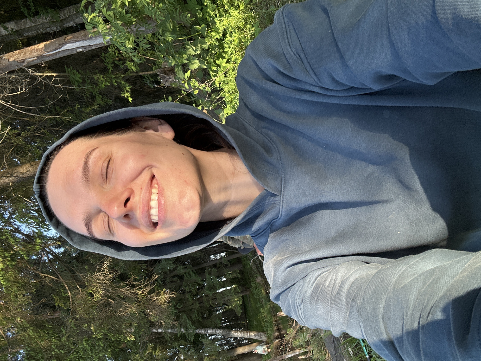 A woman with closed eyes and a wide smile, wearing a blue hooded sweatshirt, takes a selfie outdoors. She appears to be standing in a forested area, with trees and greenery visible in the background. The location is unknown, but is specified as Bormist, Russia.