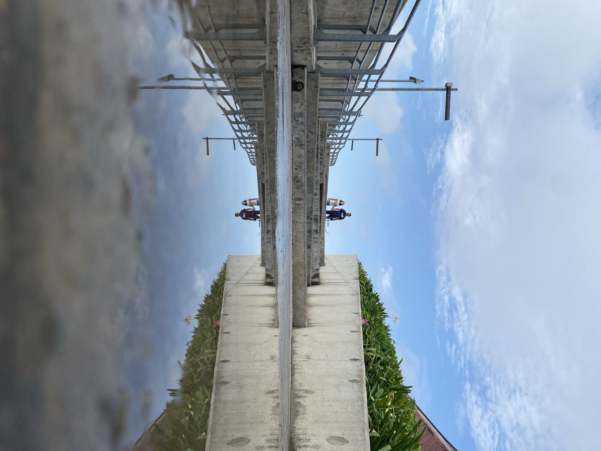 An upside-down medium shot presents an elevated pedestrian walkway or bridge in Buenos Aires, Argentina. The bridge's concrete supports are adorned with dense green vegetation. Two people are positioned at the bridge's midpoint, seemingly looking out. The sky, with scattered clouds, reflects beneath the structure, creating a mirrored effect. The bridge's framework is constructed from gray metal.