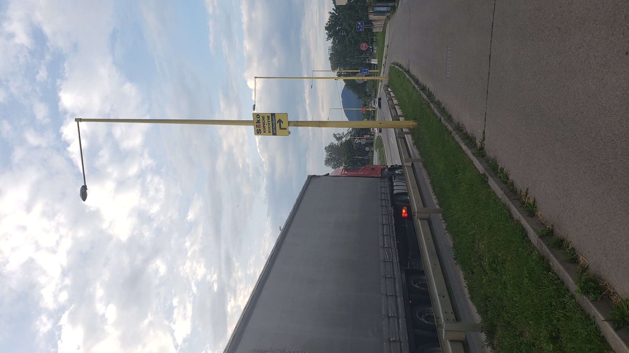 The image captures a roadside scene in Prešov, Slovakia, on a bright, partly cloudy day. The perspective is from alongside a road, with a large, red and grey semi-trailer truck prominently positioned on the right side of the frame. The truck appears to be traveling or stopped alongside a metal guardrail and a strip of green grass.
To the left of the truck, several tall, yellow utility poles line the road. One of these poles displays a yellow rectangular sign with black text and an arrow. The text on the sign reads: "SIKO", "KÚPEĽNE" (Bathrooms), "KUCHYNE" (Kitchens), and "pri Hornbachu" (near Hornbach), with an arrow pointing to the left, indicating the direction for these businesses. A streetlight fixture hangs from another yellow pole, extending over what would be the road.
The main asphalt road stretches into the background, where more trees, other vehicles (including a car and another truck), and a distant mountain or large hill are visible under the expansive sky. The sky itself shows a mix of bright blue patches and scattered white clouds. The overall scene depicts a typical highway or major road environment in an urban or suburban area.