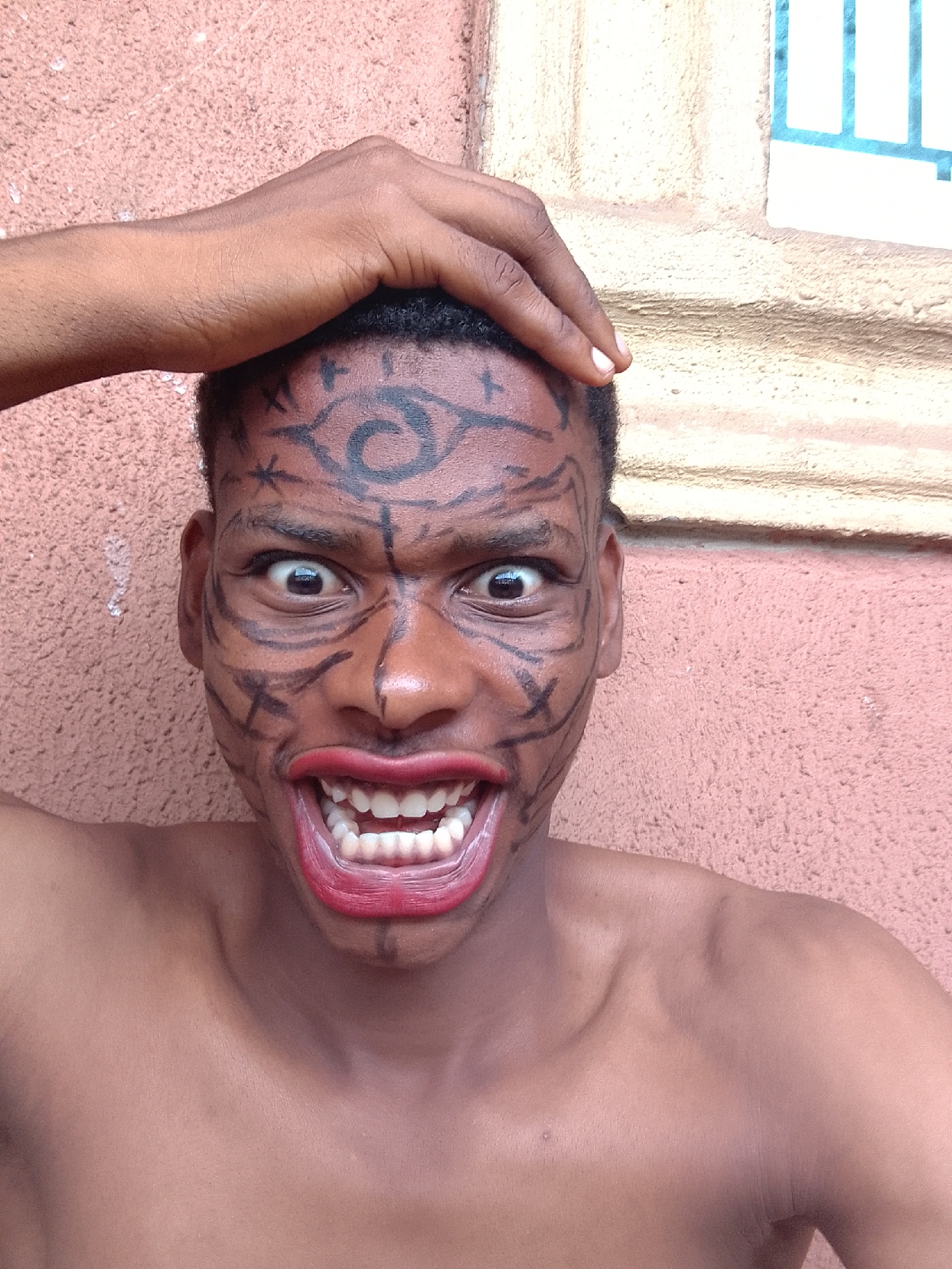 A close-up photograph shows a dark-skinned person with black face paint arranged in geometric patterns and a spiral design on the forehead, bearing a resemblance to symbols from the Naruto anime series. The person has wide eyes and is baring their teeth, with red lipstick applied. Their hand is resting on their head. The subject appears to be shirtless. Behind the person is a textured pink or light brown wall, with a window partially visible in the upper right corner. The location is specified as Unknown, Unknown.