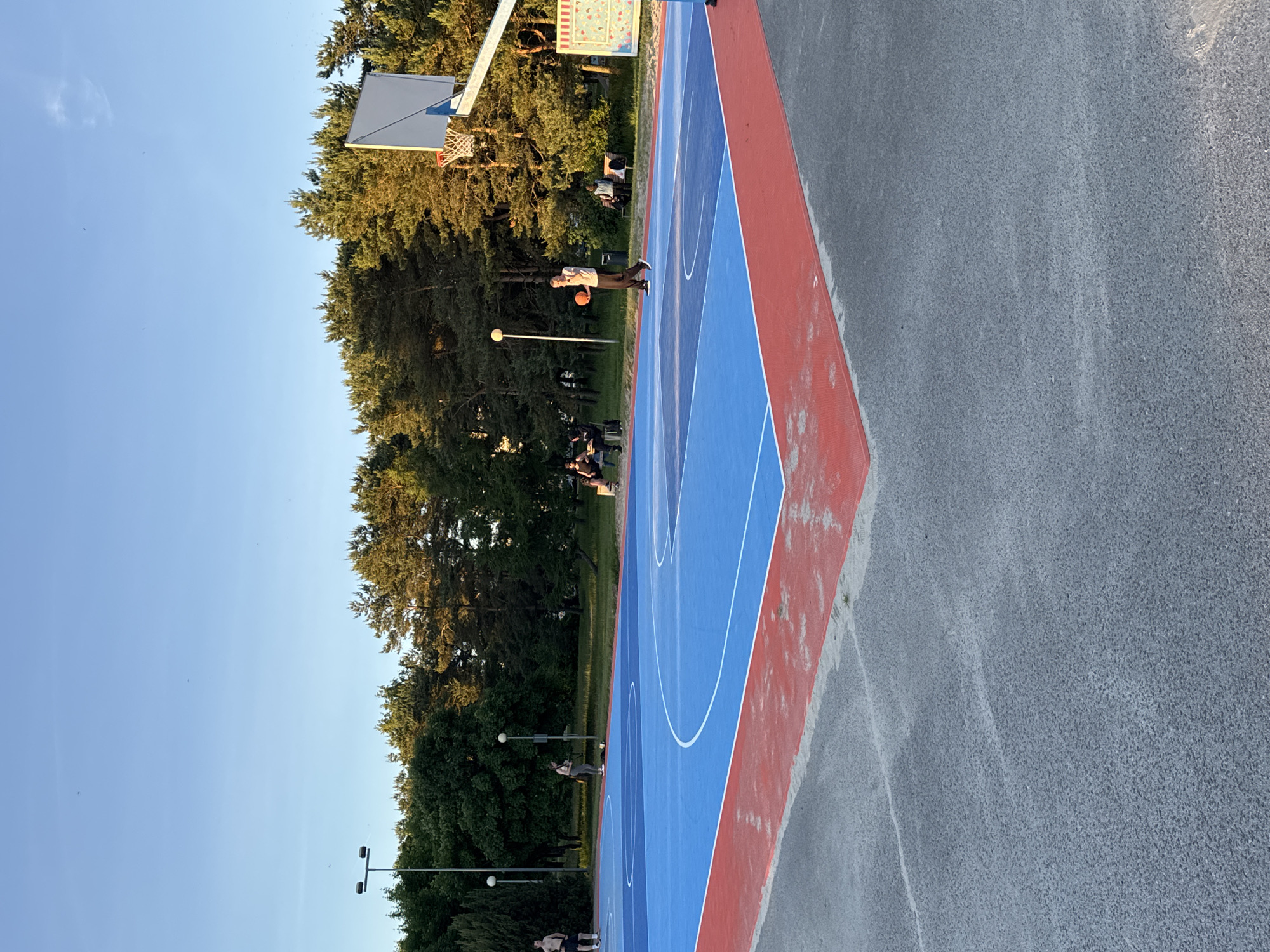 The image shows an outdoor basketball court with a blue and red surface. The court is lined with white markings. Several people are present on and around the court. One man is dribbling a basketball on the court. Others are sitting on benches in the background. Trees and a clear blue sky are visible behind the court. The scene appears to be in a park or recreational area. The style of the basketball court, with its bright colors and surrounding parkland, is consistent with public sports facilities found in cities like Tallinn, Estonia.