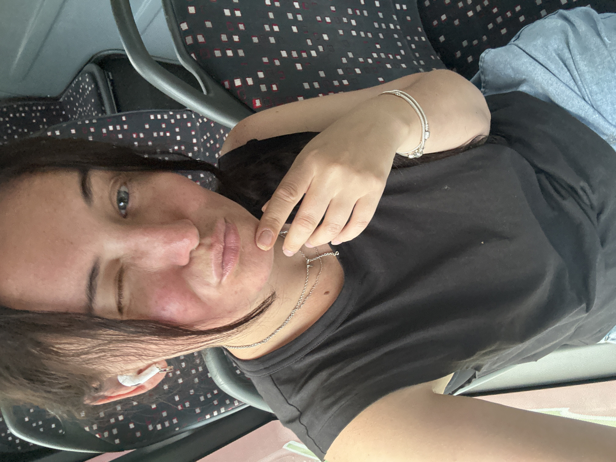 A woman with dark hair and fair skin is taking a selfie on what appears to be a bus or train. She is wearing a black tank top, a silver necklace, and a silver bracelet. She also has white earbuds in her ears and is winking at the camera. The woman's face is slightly red, possibly from blushing or sun exposure. Behind her, the seats of the vehicle are visible, with a patterned upholstery. The lighting suggests it is daytime. The image does not provide any specific clues about the location in the city of Günzburg, Germany, or any other city.