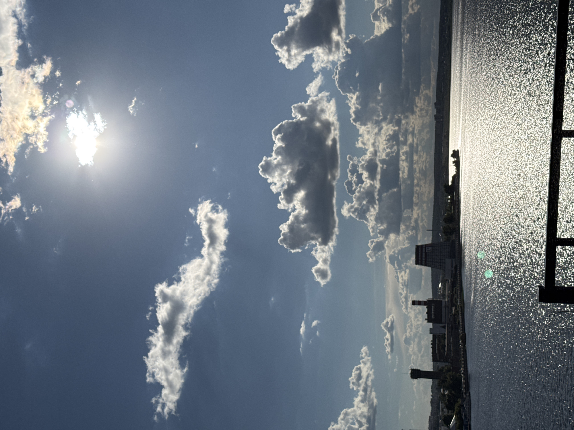 The image shows a view of the Kama River in Izhevsk, Russia, under a sky filled with clouds. The sun is visible behind some clouds, creating a bright glare. Along the riverbank, there's a cityscape with buildings and industrial structures, including a power plant with tall chimneys. The river surface is shimmering due to sunlight reflection. A partial view of a structure with vertical bars is visible at the right edge of the image.