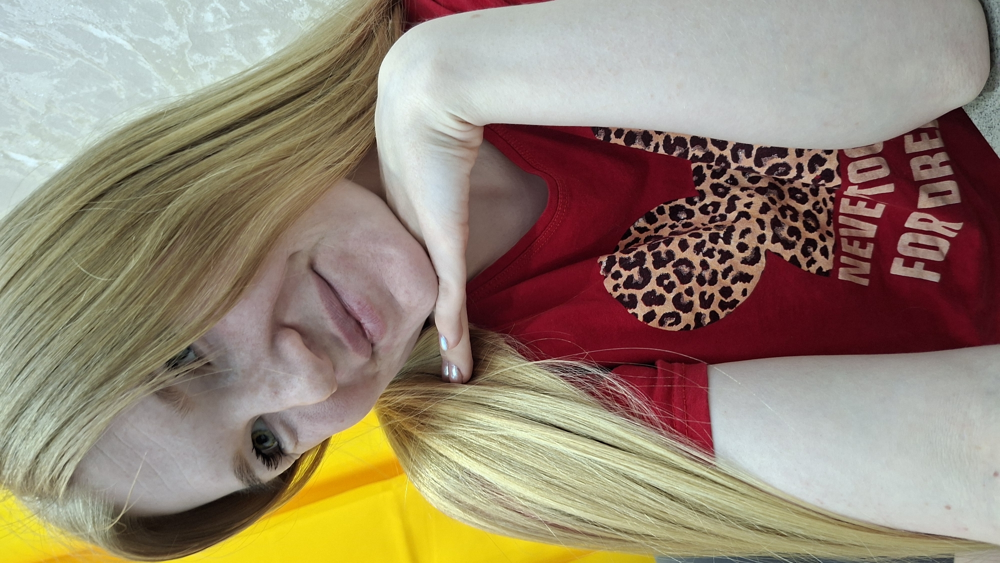 A fair-skinned woman with blonde hair poses for a selfie indoors. She is wearing a red t-shirt with a leopard-print Mickey Mouse head design and the words "Never too old for dreams." Her left hand is touching her cheek. The environment appears to be a domestic setting. There is no definitive indication that the photo was taken in Chaikovsky, Russia.