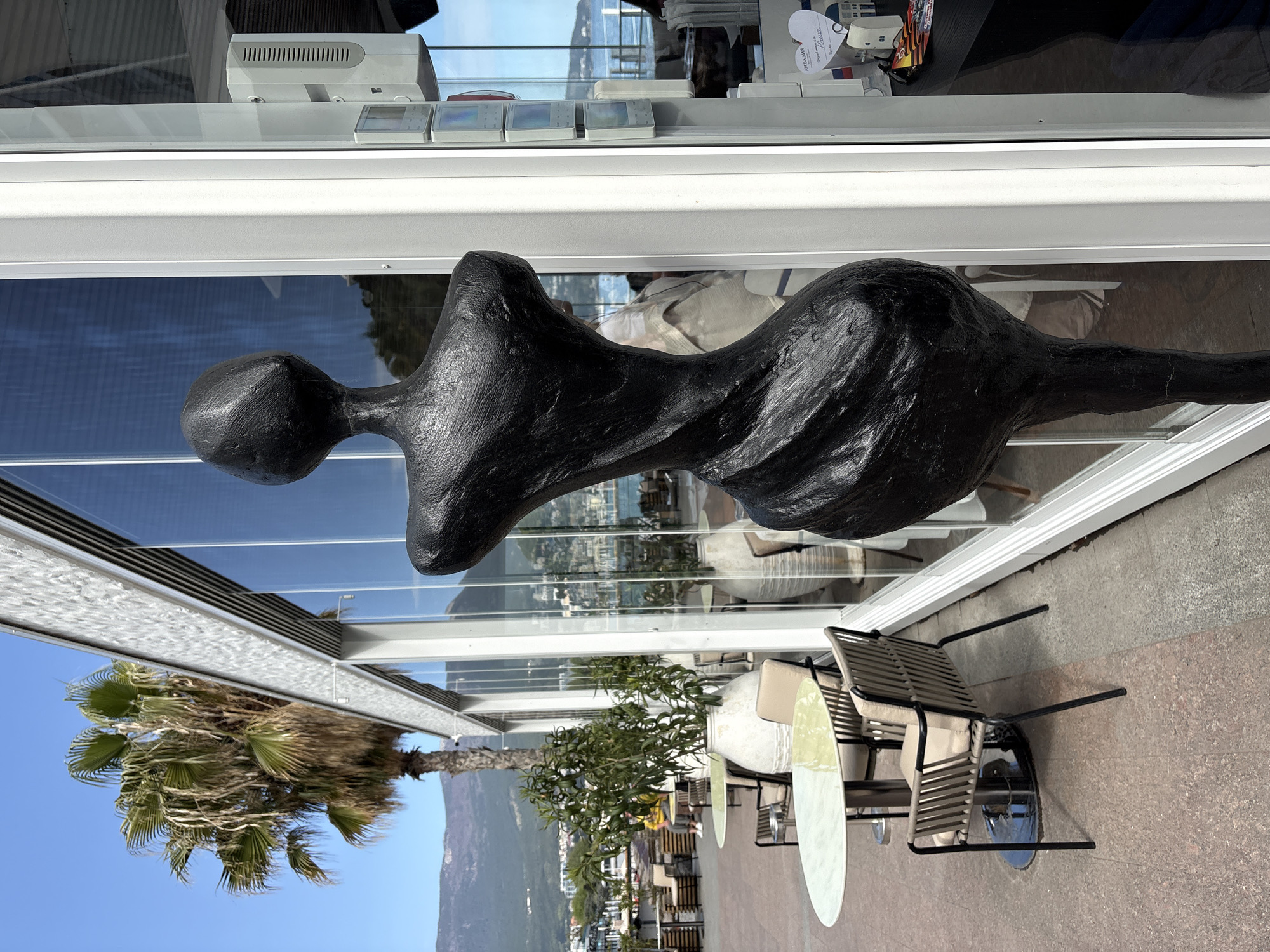 The image features an abstract sculpture of a human figure with a smooth head, a narrow neck, and a swelling torso. The sculpture is positioned inside a modern building with large glass windows, reflecting the outdoor scene. Outside the building, there are palm trees, other trees, tables, chairs, and what appears to be a cityscape with mountains in the background. The location is identified as город Ялта, Ukraine. The building seems to be a restaurant or a shop, with some objects visible behind the glass windows. There are no people visible in the outdoor scene.