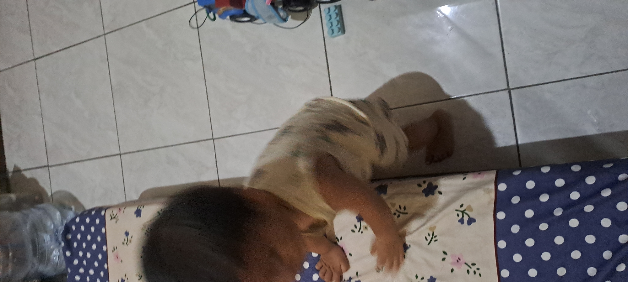 A child is bending down on a floral patterned surface, their head is dark and their clothing is light-colored and patterned. The child is on a surface that appears to be a mattress or bed. Next to the mattress is a tiled floor. In the background, there are some toys and a large water bottle. The scene takes place indoors in Cemandi, Indonesia.
