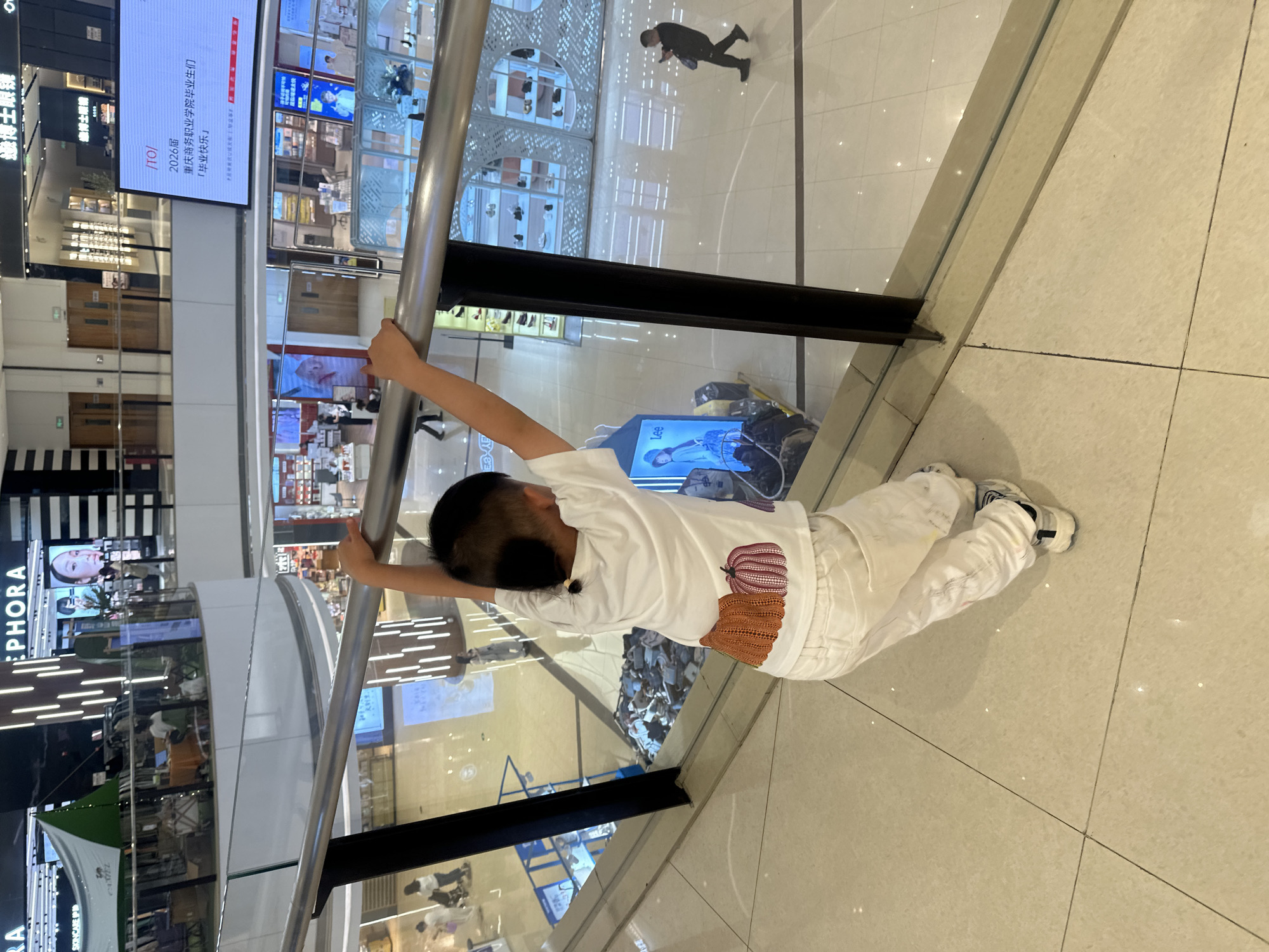 The image captures a child in a white t-shirt and white pants, with a pumpkin design on the back of the shirt, holding onto a silver railing in a modern shopping mall. The child is positioned on an upper level, looking down into the mall. In the background, stores and displays are visible, along with other shoppers. The environment appears clean and well-lit, typical of a large retail space. The presence of signs with Chinese text and specific store names like "SEPHORA" and what appears to be an electronic billboard with Chinese characters suggest the location is within China. Specifically, the electronic billboard displays text that includes "JTO/ 2026届 重庆商务职业学院毕业生们『毕业快乐』", indicating the location is in Chongqing and likely references a graduation event for Chongqing Business Vocational College. This strongly points to the scene being in the Chongqing High-tech Zone, China, a common area for commercial and educational facilities.