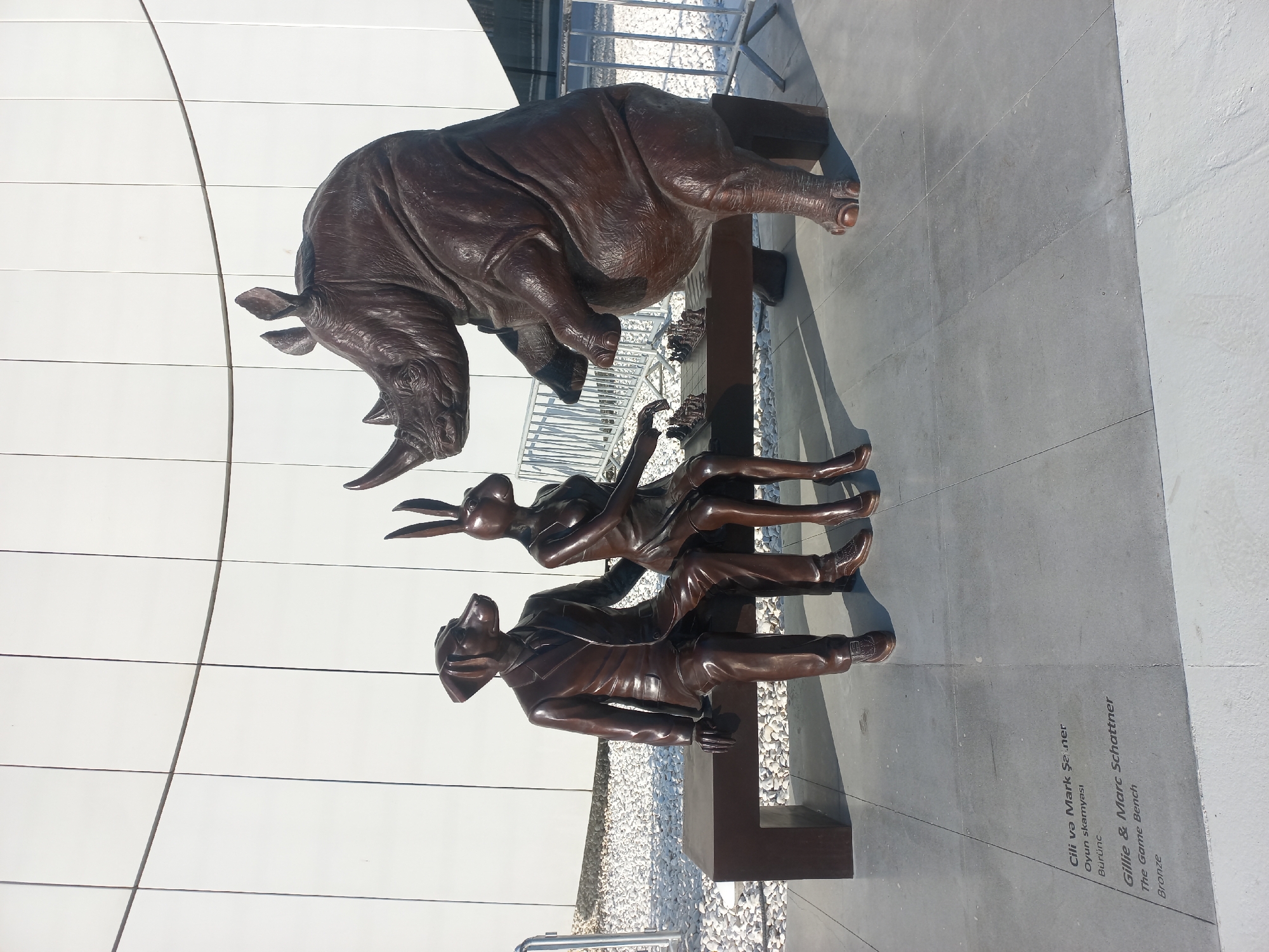 The image depicts a bronze sculpture installation titled "The Game Bench" by artists Gillie and Marc Schattner, located in Baku, Azerbaijan. The sculpture features a rhinoceros standing on its hind legs, with its front legs extended towards a chessboard set up on a bench. Seated on the bench to the left of the chessboard are two anthropomorphic figures: one resembling a dog wearing a suit and tie, and the other a rabbit wearing a dress. They are both looking towards the chessboard. The ground in front of the sculpture has inscribed text in both Azerbaijani and English identifying the artwork and artists. The background consists of a white, curved modern building facade and metal fencing. The scene appears to be outdoors on a sunny day.