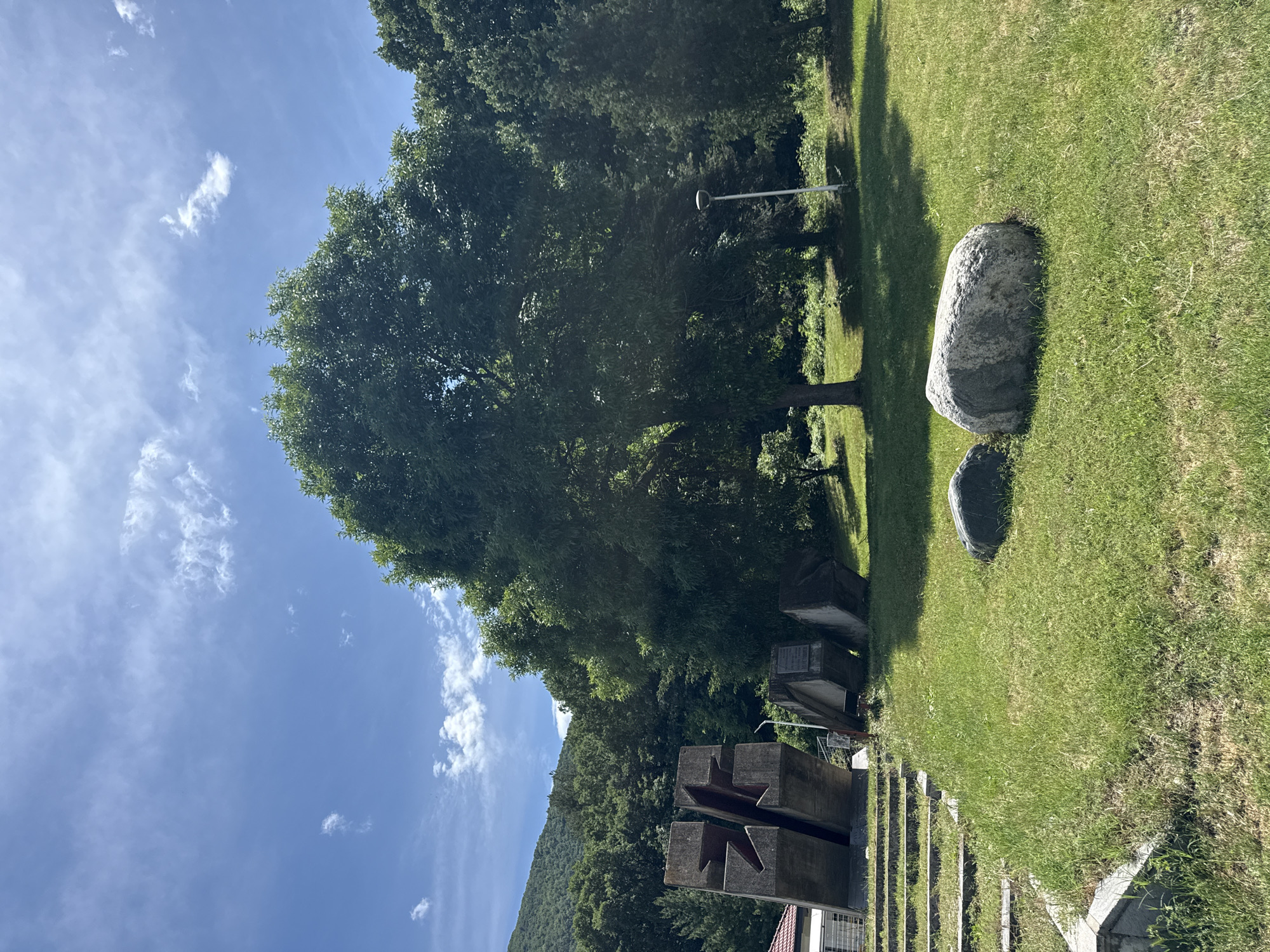 This image depicts an outdoor memorial or park area in Vetren Dol, Bulgaria, bathed in bright midday or early afternoon sunlight, evident from the strong shadows cast on the lush green grass. The sky is clear blue with scattered white, wispy clouds.
In the foreground, a large, weathered concrete monument dominates the left side. It comprises several heavy, block-like structures, two of which feature deeply carved, stylized star or cross-like shapes. A small section of a building with a reddish-orange roof is visible at the very bottom left. The ground around the monument is covered in green grass.
The midground features a sprawling green lawn where two smooth, grey boulders of differing sizes are placed. A tall, slender white pole, possibly a lamppost, stands upright among the trees. Numerous large, mature trees with dense green foliage provide shade and frame the scene, their leaves vibrant under the sun.
In the background, a dense forest covers a steep hillside or mountain, extending under the clear sky. No people or specific activities are visible, lending a tranquil and contemplative atmosphere to the scene. While there appears to be a plaque or inscription on one of the concrete monument blocks, the text is not legible due to the distance and resolution.