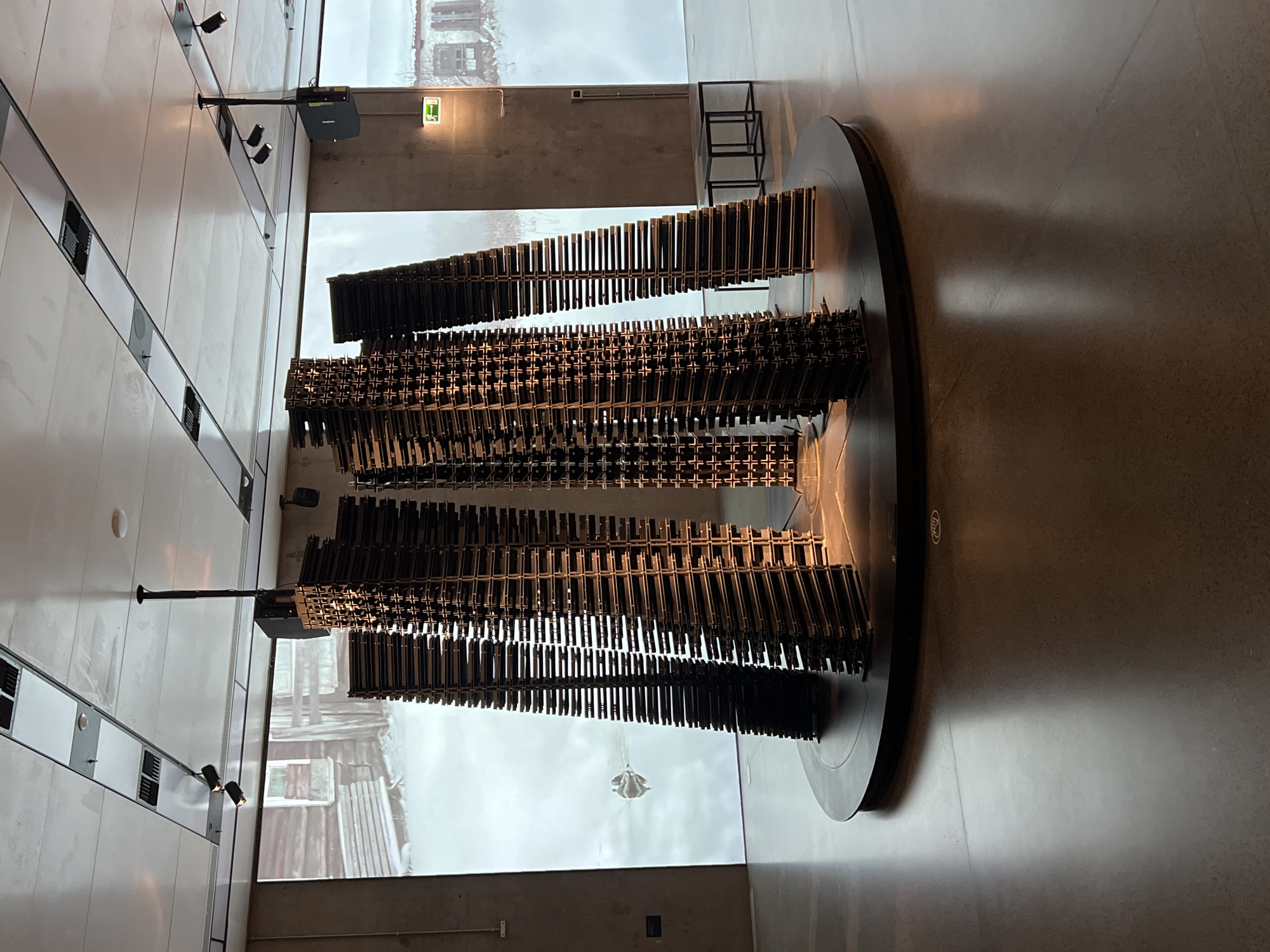 The image depicts an art installation inside a museum. The installation features numerous metal chairs arranged in several columns on a large, circular platform. Behind the sculpture is a projection on a large screen that shows a black and white image of what appears to be a building. The background of the image is the museum's architecture: a pale tiled wall with linear vents, and a concrete wall with a green emergency exit sign. A spotlighting system is attached to the ceiling. The location is Unknown, Unknown.