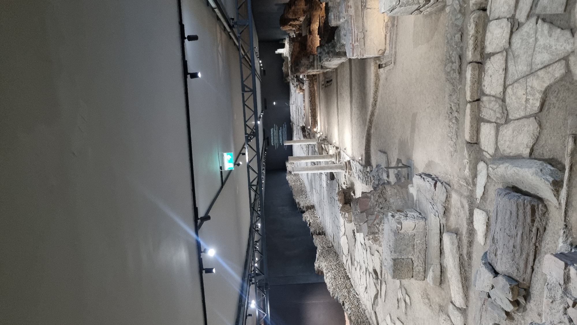 The image depicts the archaeological site of the Roman Agora in Thessaloniki, Greece. The scene is an interior view of an excavated ancient Roman complex, now preserved and displayed indoors. The environment consists of stone ruins, including paved pathways, foundations of buildings, partial walls, and several large stone columns. The ruins are lit by artificial lighting, with spotlights casting illumination on various sections of the site. In the background, a dark wall with projected text or images is visible, suggesting an informational display. Above, the ceiling features a grid of track lighting. No people are present in the image. The overall impression is one of a preserved historical site designed for public viewing and education.