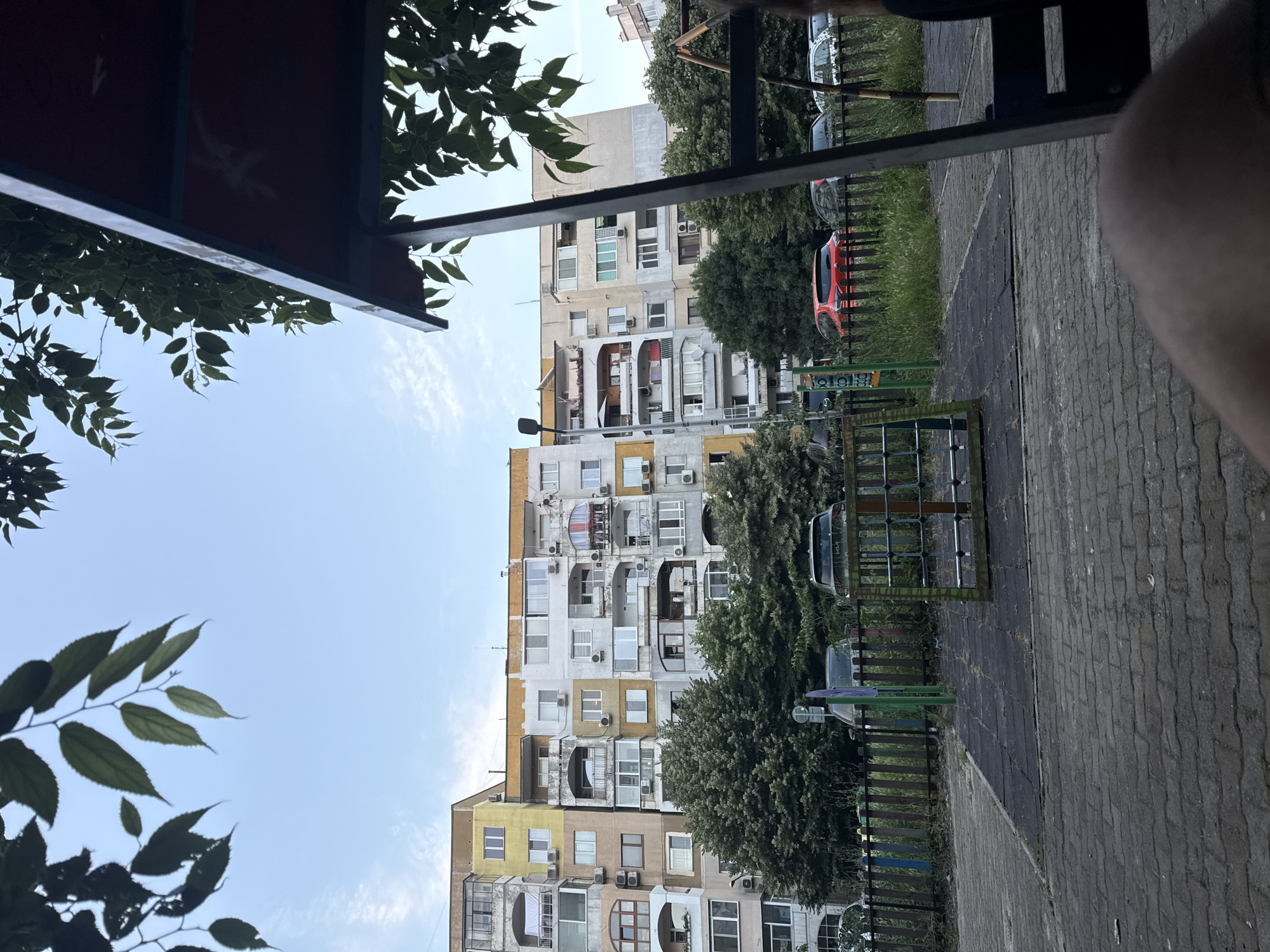 The image shows an outdoor scene in Varna, Bulgaria, with a residential building in the background and a playground in the foreground. The building is multi-story and appears to be an apartment complex, with a mix of yellow, white, and grey facades. Many windows and balconies are visible, some with air conditioning units. Trees and foliage are present in front of the building and along the perimeter of the playground.
The playground area includes a rubberized surface and a small climbing structure made of metal and rope. A wooden fence, painted in various colors, separates the playground from a grassy area where several cars are parked. One car is bright red, and others are dark-colored. The sky is clear with some light clouds. The image is taken from a low angle, possibly from a seated position, with part of a person's leg or arm visible in the bottom right corner. The overall impression is a common urban scene in a residential area.