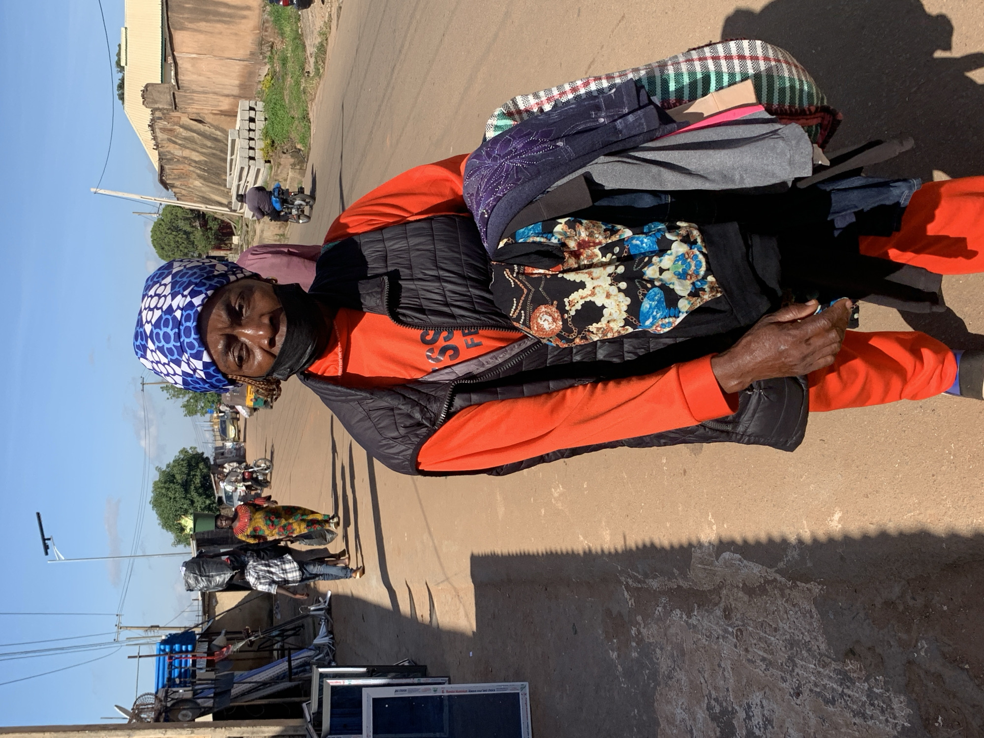 In Gura Top, Nigeria, a woman wearing an orange long-sleeved shirt, a black vest, and an orange headscarf walks along a dirt road. She is carrying a bundle of clothes in her arms, including a black patterned fabric with floral designs, a dark blue patterned scarf, and a plaid fabric. She also wears a black face mask. The street is lined with shops and buildings, and other people are visible in the background, including a person on a motorcycle and a woman carrying a large bag on her head. The sky is clear and blue.