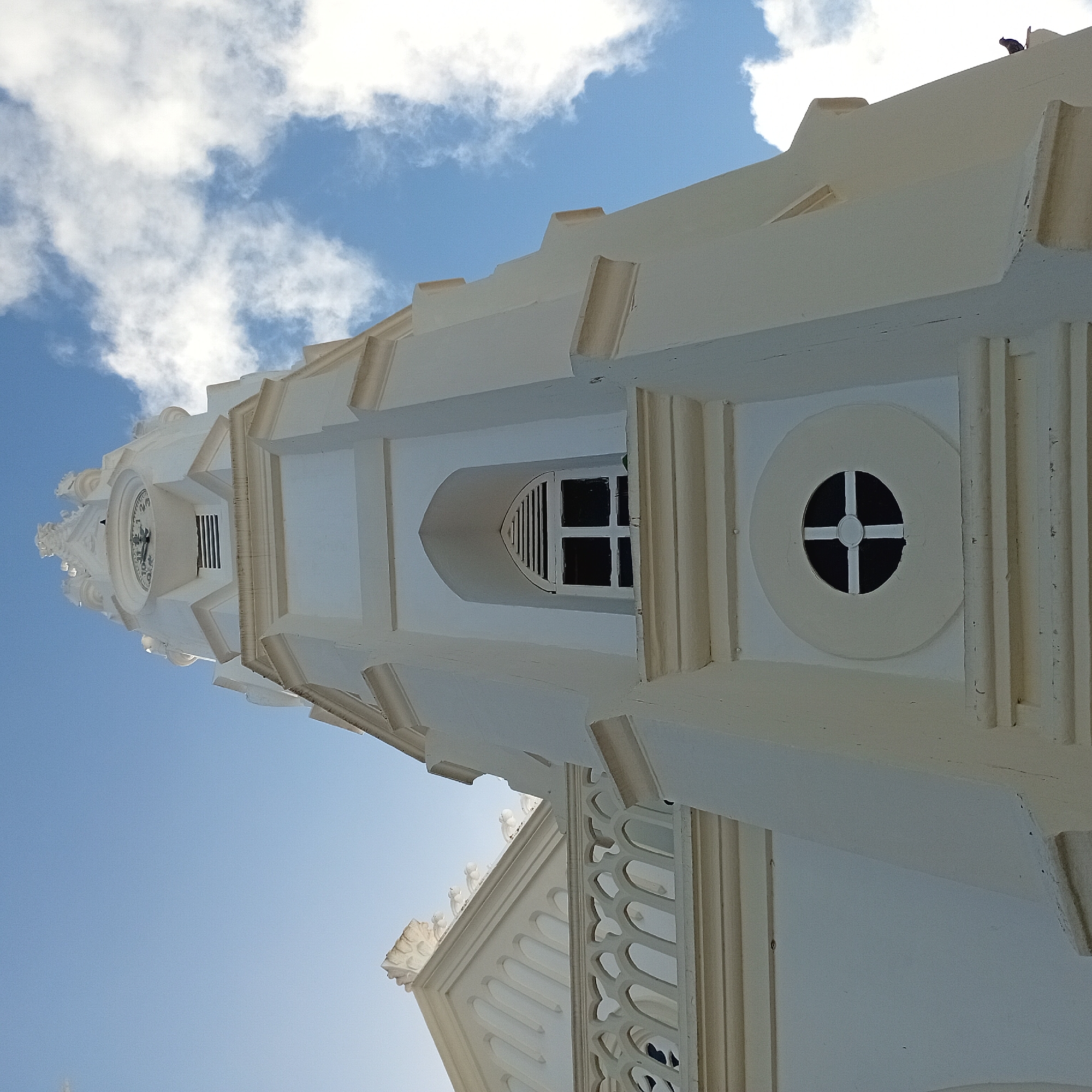 The image shows a low-angle view of a white building's tower against a blue sky with some clouds. The tower features architectural details such as a clock face, various windows (including one with a cross pattern), and decorative molding. A bird is perched on top of the building, near the upper right corner of the image. The building appears to be old with traditional design elements. The location is unknown.