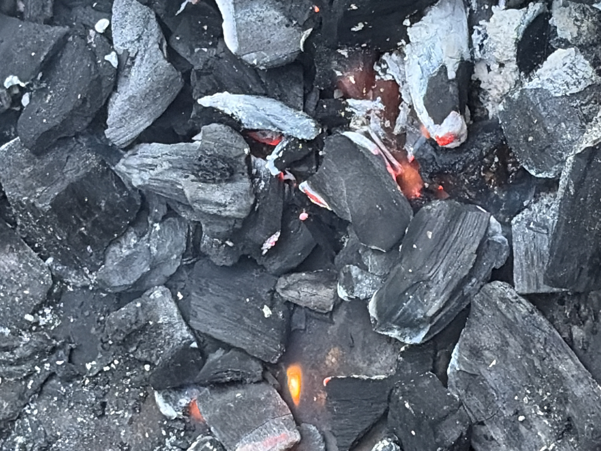 A close-up, overhead shot captures a pile of smoldering charcoal. The charcoal pieces are irregularly shaped, varying in size from small fragments to larger chunks, with a rough, porous texture. Some pieces are uniformly dark gray, while others show hints of glowing orange and red where they are actively burning, emitting a soft light. White ash dust is visible on some of the charcoal surfaces and scattered between them. The lighting is dim, with the primary light source appearing to be the heat from the coals, casting subtle shadows. The background is filled with more charcoal, creating a dense, textured landscape. The scene is devoid of any human presence or identifiable urban features, making it impossible to determine a specific location in Snegiryovka, Russia, or any other city.