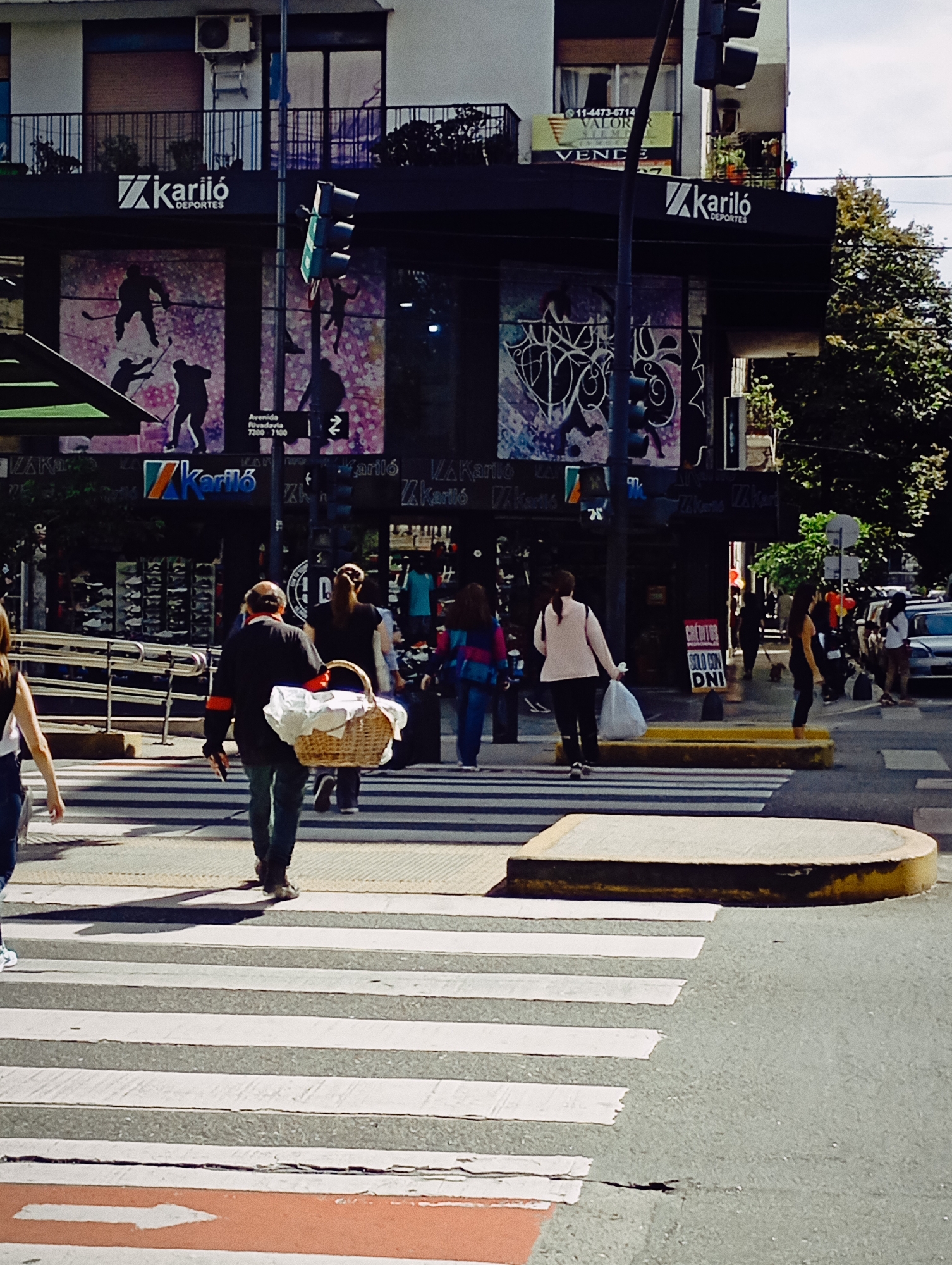 The image shows a street scene in Buenos Aires, Argentina, focusing on a pedestrian crosswalk in front of a "Karilo Deportes" store. People are walking across the crosswalk, including a man carrying a basket. The crosswalk features white stripes on the pavement, with a red section on one end. A traffic light pole is visible near the store, and other buildings, trees, and vehicles appear in the background. The "Karilo Deportes" store has advertisements and signage, including store hours "Avenida Rivadavia 7200-7100". Other signs read "SE VENDE" and "Créditos solo con DNI."