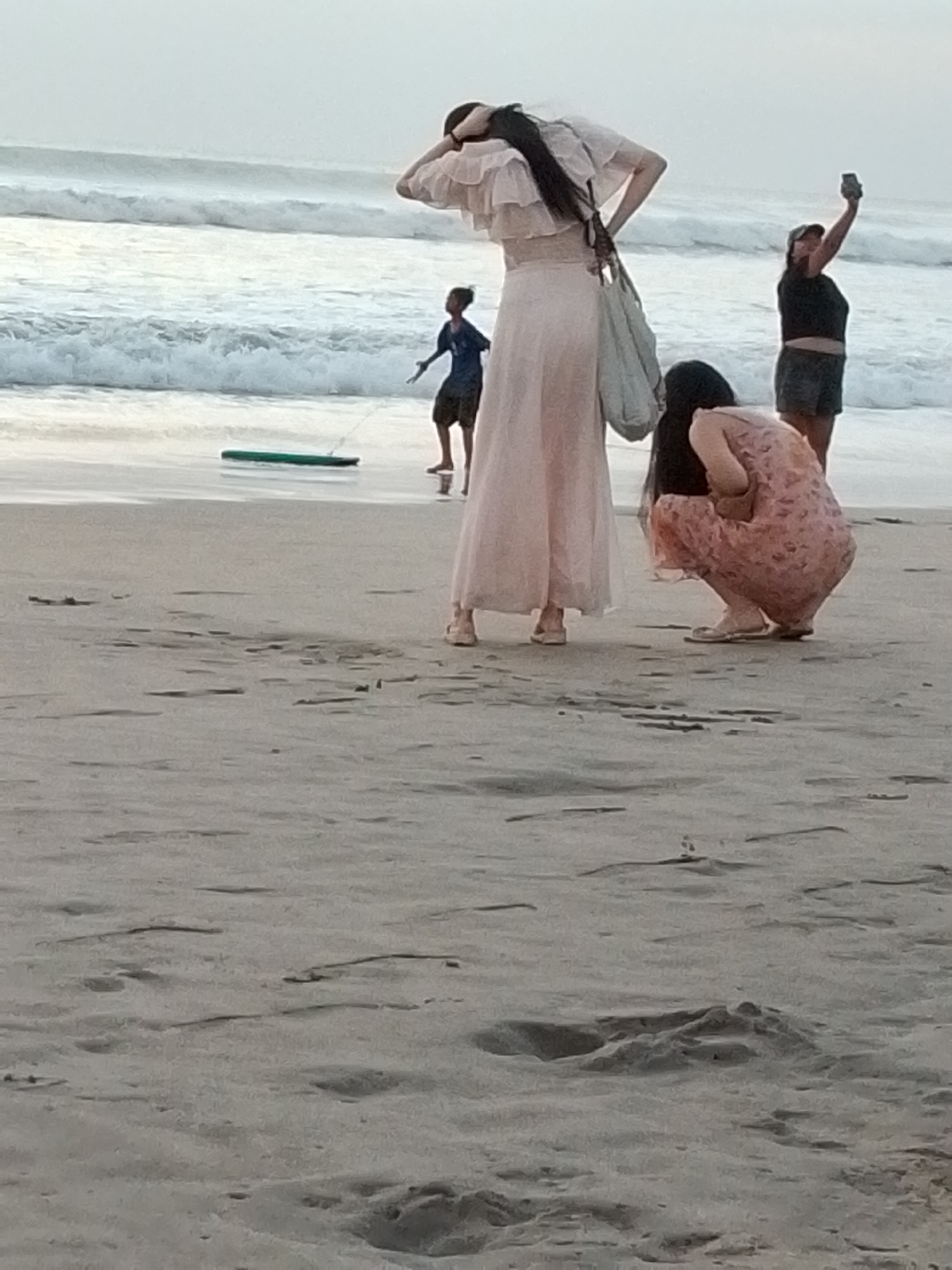 In Kuta, Indonesia, four individuals are observed on a sandy beach with waves in the background. The scene is captured in an outdoor, natural light setting.
A woman in the foreground is crouching down, facing away from the camera, wearing a pink floral dress and sandals. To her left and slightly behind her, another woman stands tall, wearing a long, light pink dress with ruffled sleeves and sandals. She has long dark hair and is holding a light-colored bag.
Further in the background, a person in a dark blue t-shirt and shorts stands near the water's edge, facing the waves and holding a green boogie board connected by a string. To the far right, a woman in a black t-shirt and dark shorts is standing and holding up a mobile phone, possibly taking a picture or video. The beach is covered in sand with visible footprints. The ocean has moderate waves.