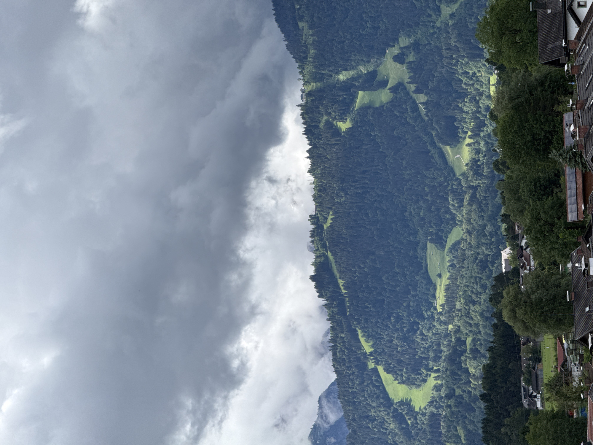 This media file depicts a panoramic view of a mountainous landscape in Garmisch-Partenkirchen, Germany. The scene is dominated by a steep, densely forested mountainside covered in dark green pine trees. Patches of bright green grass are visible on the slopes, suggesting clearings or meadows. A winding path or road can be seen snaking its way through the trees.
On the right side of the image, a small village is nestled along the base of the mountain. Several houses with dark, sloped roofs are visible, interspersed with trees. The architecture appears to be traditional, with some buildings featuring multiple stories and visible windows.
The sky on the left side of the image is filled with dramatic, heavy, gray clouds, indicating overcast or potentially stormy weather. The contrast between the dark, moody sky and the vibrant green of the mountain and meadows is striking. There is no visible text in the image. The time of day is not definitively clear due to the cloud cover, but it appears to be daytime. The overall mood is one of natural grandeur and a sense of the vastness of the mountain environment, with human habitation present but small in scale compared to the surrounding nature.