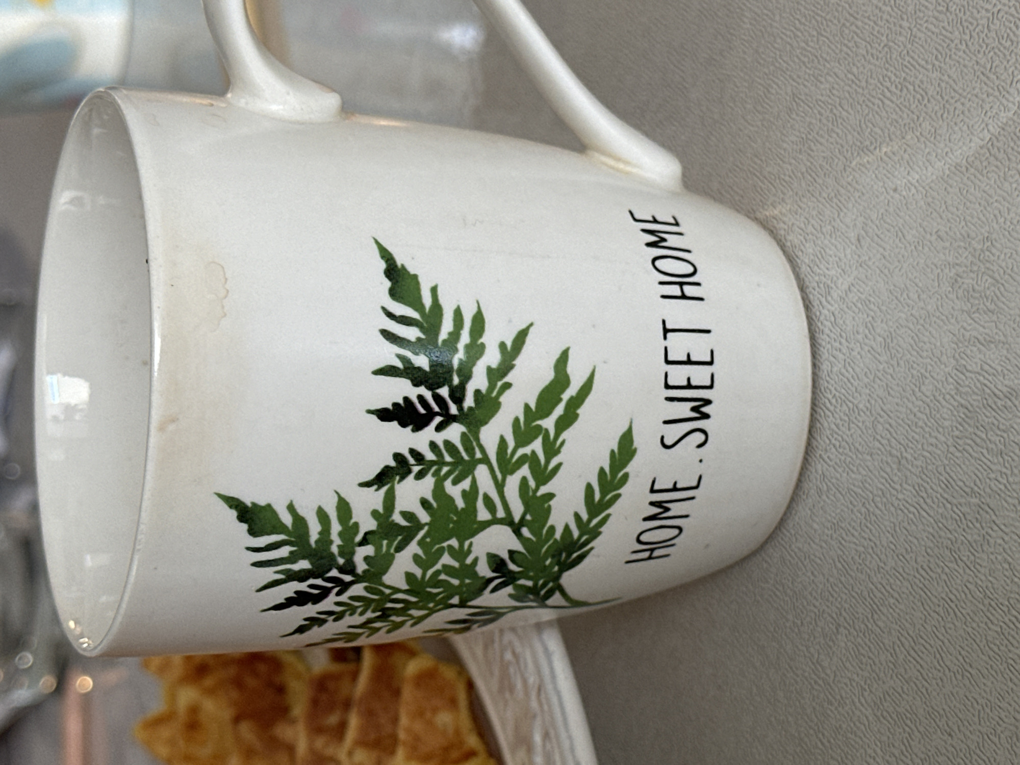 A close-up, eye-level shot focuses on a white ceramic mug with a black fern decal and the words "HOME SWEET HOME" printed in black lettering. The mug has a handle and a slight brown discoloration near the rim. To the left of the mug, partially visible, are several slices of golden-brown waffles stacked on a white plate. The mug and plate rest on a textured gray surface. The background is blurred, showing hints of other objects and a light source. The scene appears to be set indoors, on a table. The image does not contain any recognizable landmarks or clues that would indicate its location in Рабочий, Russia.