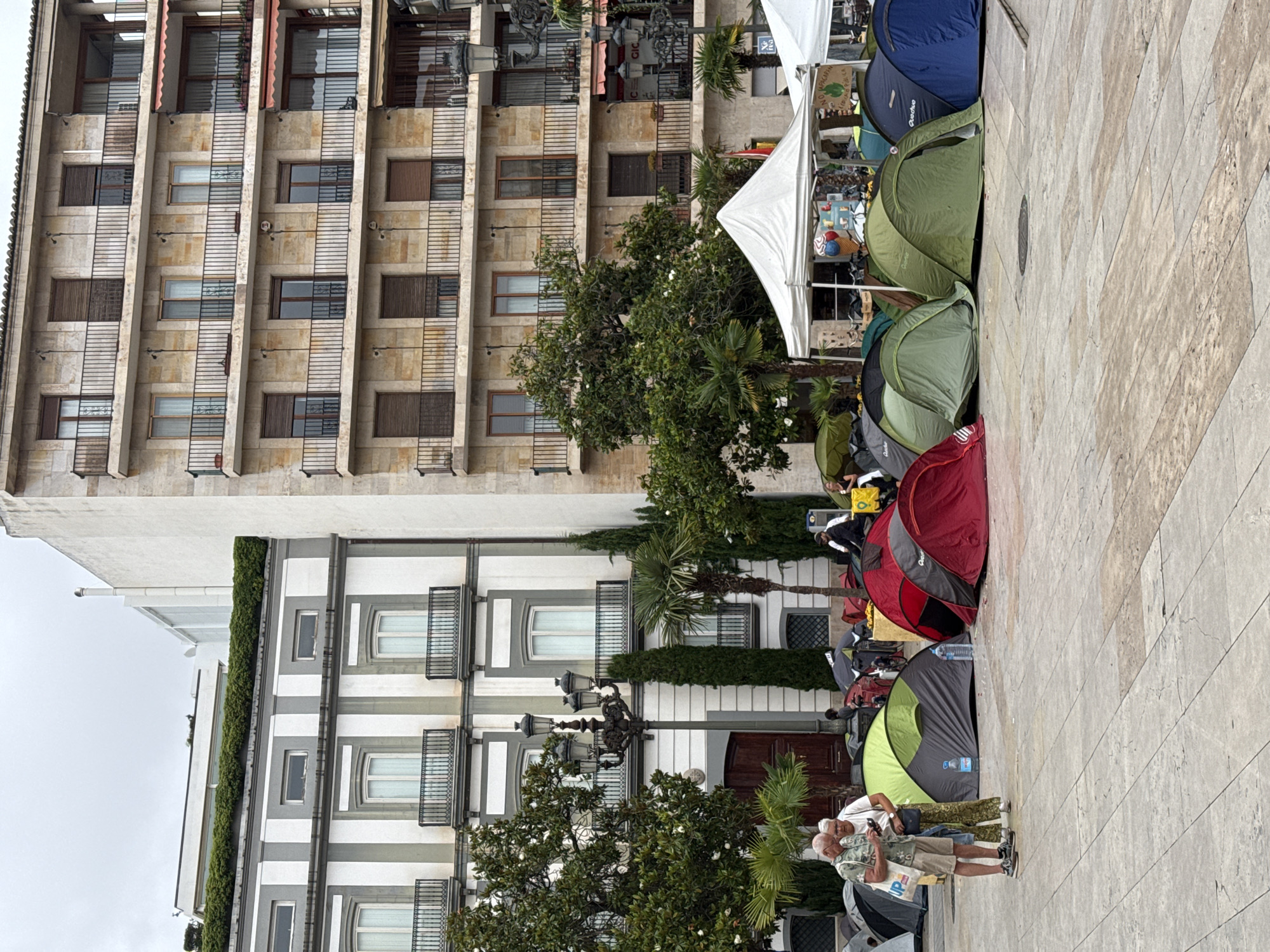 This image depicts an urban outdoor scene in València, Spain, featuring a gathering of tents and people on a wide paved plaza. The foreground is dominated by a row of colorful pop-up tents, primarily green, blue, red, and a grey and lime green variation. Many of these tents bear the "Quechua" brand. Several white canopies are also set up, providing shelter or serving as temporary stalls.
People are visible around the tents and canopies. In the immediate foreground, a man wearing a camouflage-patterned shirt and beige shorts stands next to a tent, looking at a device. Nearby, a blue plastic bottle with the label "FONT SALEM" is visible. Under one white canopy, a green sign reads "NO A LA MACROPLANTA... PÚBLIC," suggesting a protest or demonstration against a large industrial plant and advocating for public interests.
The background features several multi-story buildings. One building, directly behind the tents, has a light brown stone facade with numerous windows, balconies, and horizontal metal grilles. To the left, another building exhibits a distinct architectural style with white and grey vertical stripes, large windows, and ornate iron balconies. Lush green trees, including palm trees, are scattered throughout the scene, interspersed between the buildings and the tents. An ornate black lamppost stands near the striped building.
The scene takes place during the daytime under an overcast sky, creating soft, diffused lighting. The ground is paved with large, light-colored flagstones. The overall impression is that of a temporary encampment or a long-term public gathering, possibly a protest, set within a bustling city environment.