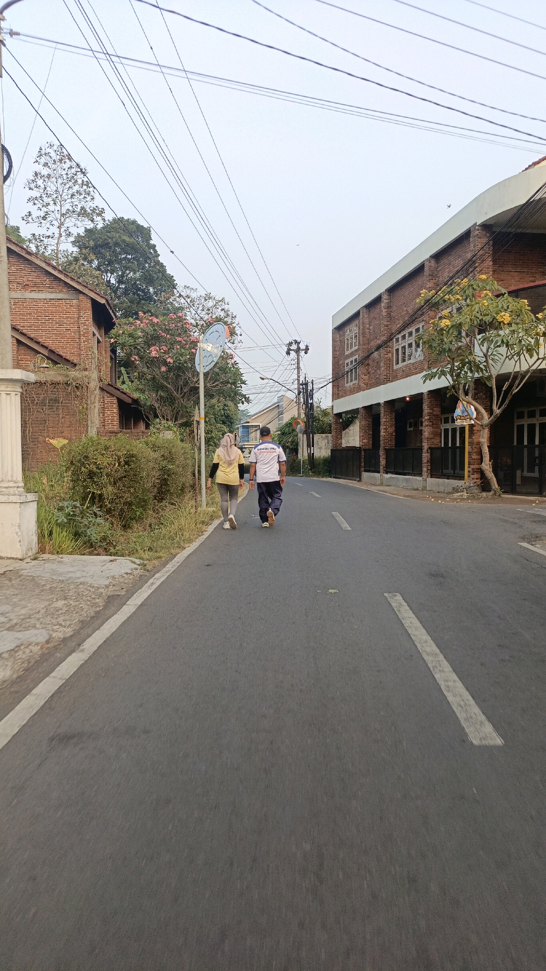 Two individuals, a woman wearing a yellow hijab and a woman in a yellow t-shirt and grey leggings, walk away from the camera on a paved road. The woman in the hijab is in front, followed by the woman in the t-shirt, who is wearing dark-colored pants and a white and blue t-shirt with text. They are walking on the right side of the road, which has a white dashed line marking its center.
The scene appears to be in a residential area of Salatiga, Indonesia. To the left of the road is a brick building with a white pillar in the foreground and overgrown greenery. Further back on the left, there is another brick building with a sloped roof and pink flowering trees. To the right of the road is a larger brick building with multiple windows on its upper floors and a covered walkway with supporting columns on the ground floor. A tree with yellow flowers is situated in front of this building. Overhead, a tangle of electrical wires stretches across the sky, and a traffic sign is visible on a pole near the pink flowering trees. The sky is overcast and a pale grey, suggesting it may be early morning or late afternoon. The overall impression is of a quiet, everyday scene in a city neighborhood.