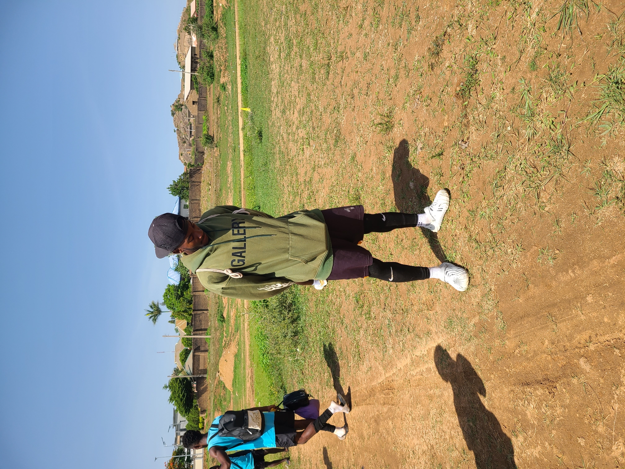The image depicts an outdoor scene in what is identified as Barikin Jirga Summa, Nigeria. The primary subject is a young male standing in the foreground, facing downwards, and wearing a light green hoodie with the word "GALLERY" printed on it in white lettering. He also wears dark shorts over black leggings, white socks, and white athletic shoes. He has a black baseball cap on his head. A white drawstring is visible hanging from his hoodie. To the left, another young male is walking away from the camera, wearing a bright blue t-shirt, black shorts, black socks, sandals, and carrying a black backpack and a purple bag. In the background, there are several structures, including brick walls, houses with varying roof types, and some hills covered with vegetation. The ground is a mixture of dirt and sparse grass. The sky is clear and blue. The overall impression is a casual, everyday scene in a semi-rural or developing urban environment.