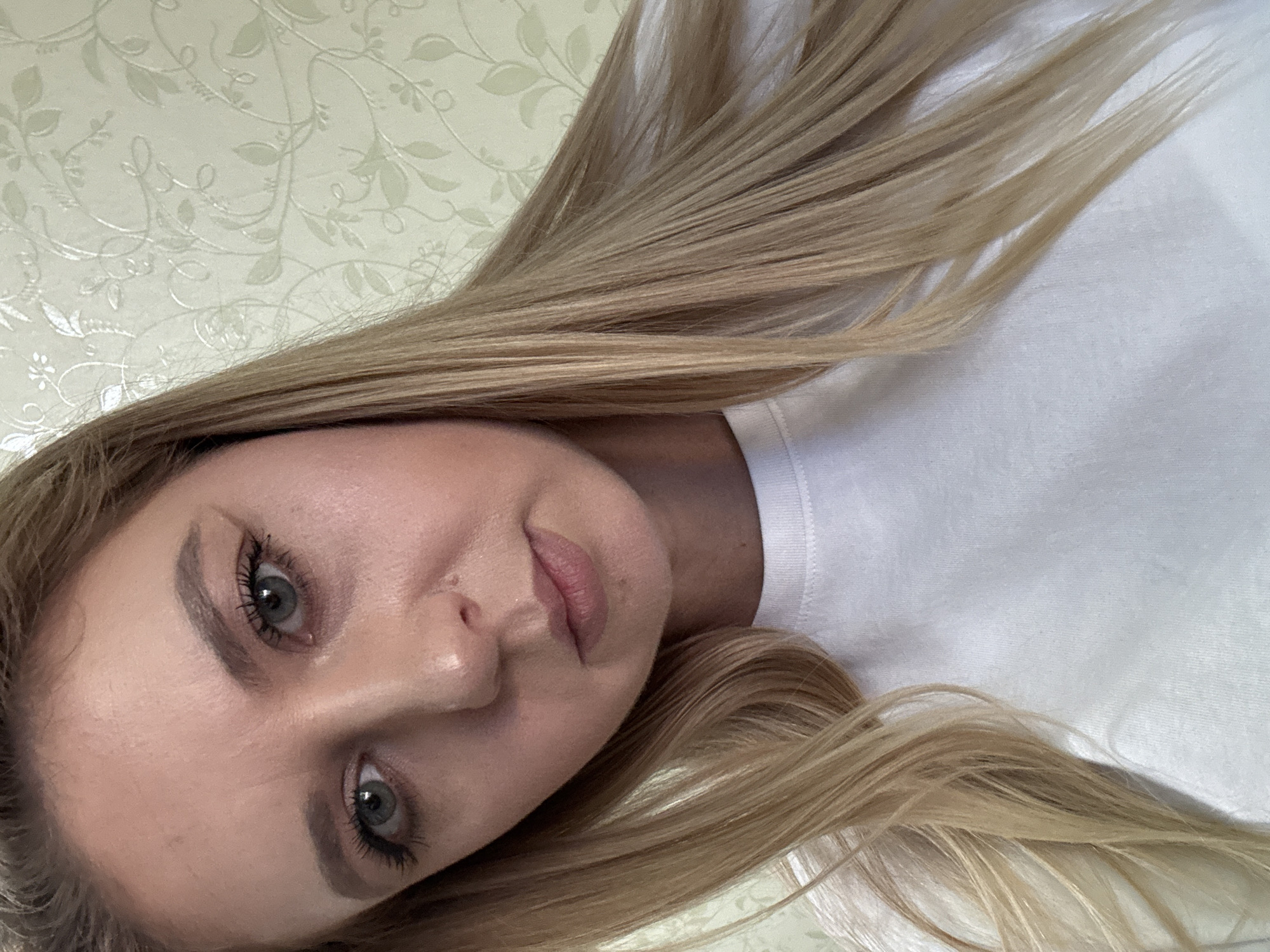 A medium shot of a woman with blonde hair and blue eyes is shown from the chest up, looking directly at the camera. She is wearing a white t-shirt. The background is a cream-colored wallpaper with a subtle floral pattern. The lighting appears to be natural.