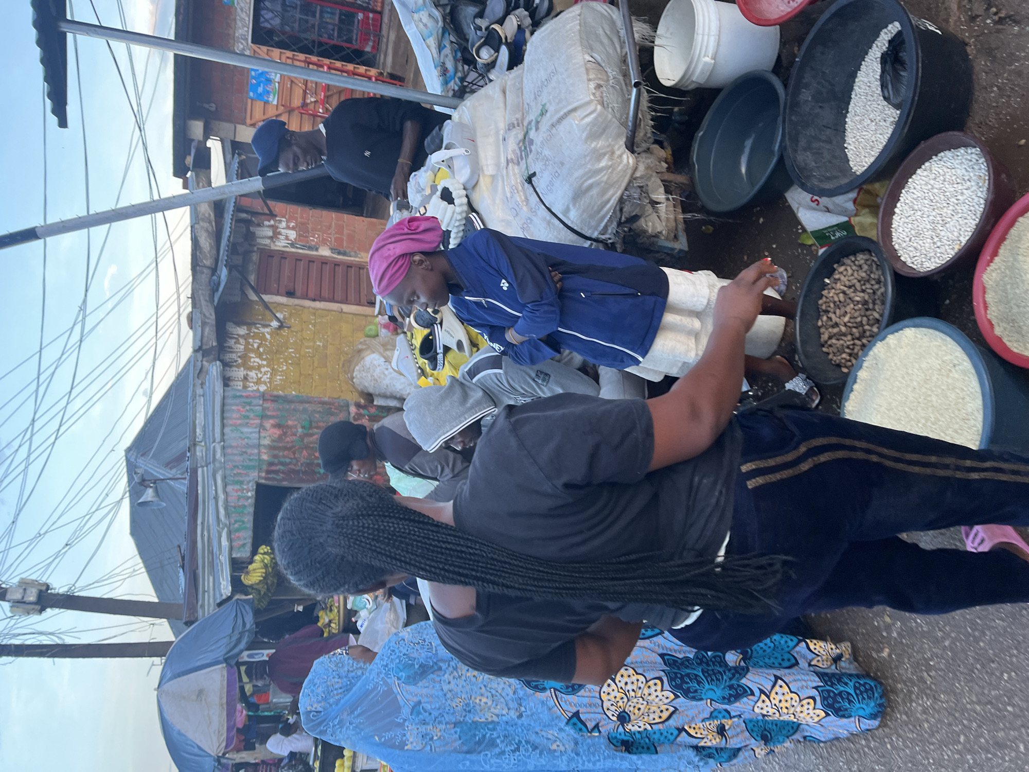 The image depicts a street scene in Gura Top, Nigeria, likely a market. In the foreground, a person with long braided hair wearing a black t-shirt and dark pants with gold stripes stands facing away from the camera. To their left, a person in a vibrant blue and yellow patterned abaya is partially visible. A young girl, wearing a blue jacket and a pink headscarf, stands to the right, looking to her left with her arms crossed. In the background, several stalls are set up with various goods. One stall displays what appears to be shoes. Another stall has sacks of produce or grains. A man in a black long-sleeved shirt and a blue baseball cap stands behind a stall. The environment appears to be an outdoor market with a rustic setup, with corrugated metal structures and electrical wires overhead. Various large plastic basins filled with grains or beans are visible on the ground in the lower right quadrant of the image. The lighting suggests it is either early morning or late afternoon.