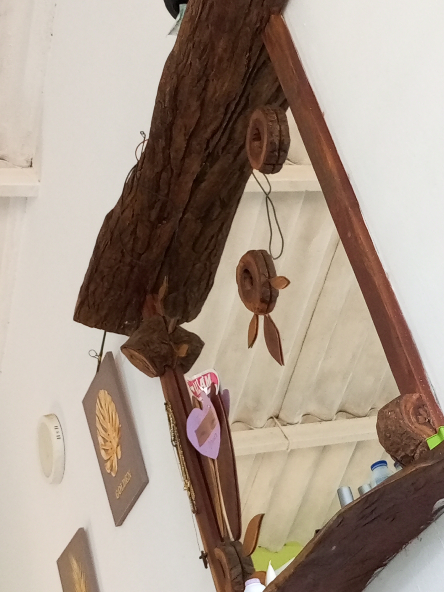 The image shows a rustic, triangular mirror with a wooden frame made of rough-hewn logs and branches. The mirror is decorated with several wooden ornaments, including two round, donut-shaped pieces with what appear to be attached wooden ears and legs, resembling small creatures. One of these hangs from a dark string. To the left of the mirror, a light-colored, oval object with vertical markings is attached to a white wall. Below this, another framed item with a golden leaf design and the word "GOLDEN" is also affixed to the wall. Further down and to the left, the corner of another similar framed item is visible. The mirror reflects the interior of a room with a white, sloped ceiling composed of parallel panels. On the shelf of the mirror's frame, to the right side of the reflection, a blue-capped bottle and some metallic cylindrical objects are visible. The overall impression is of a handcrafted, decorative piece in a casual setting. The location in La Tagua, Venezuela, is a town situated in the state of Aragua, known for its natural beauty and rural character, which aligns with the rustic aesthetic of the mirror.