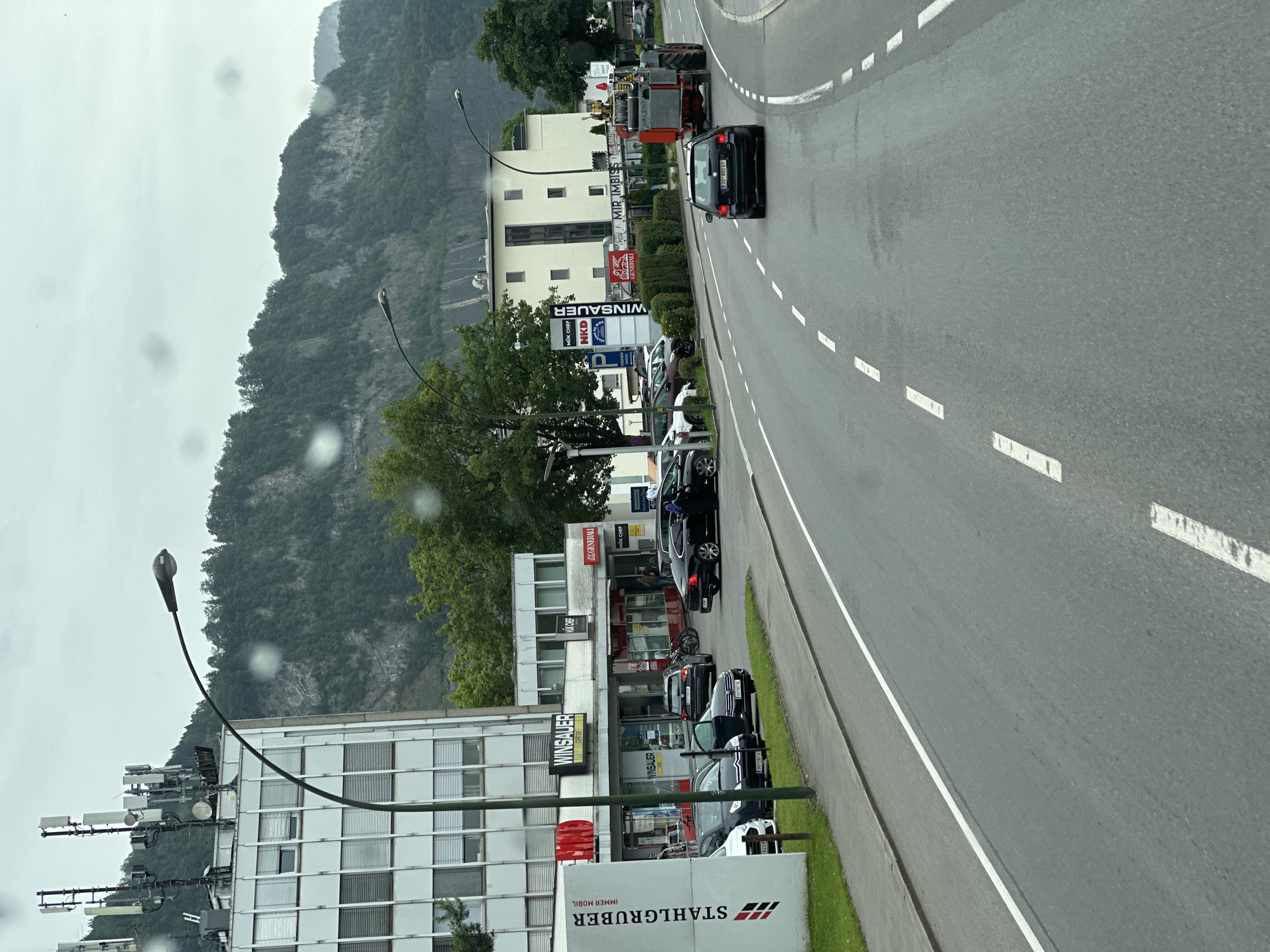 The image shows a street scene in Dornbirn, Austria, on a cloudy day. A road with lane markings runs through the center of the frame, with cars driving on it. On the left side of the road, there is a multi-story building with an advertisement for "STAHLGRUBER IMMER MOBIL" and "WINSAUER CENTER." Further down the street, there is a red and gray tractor-trailer with a water tank. In the background, there are steep, tree-covered mountains.