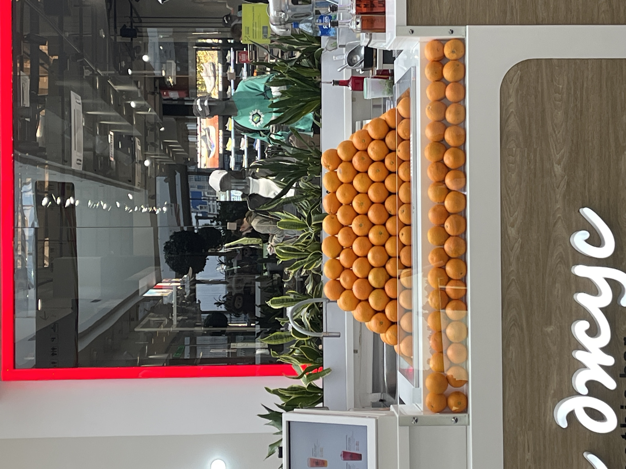 The image shows a section of a Smoothie Bar in Krasnoyarsk, Russia. The scene is captured indoors. A display case containing oranges is prominently featured; the fruits are neatly stacked in a pyramidal formation. Adjacent to the oranges is a vertical display of green plants, possibly for decoration or ingredients. Reflections of mannequins and retail displays are visible in the mirrored background, indicating the location is likely within a shopping mall or similar commercial space. The red frame on the left edge of the image may be part of a signage. To the right of the oranges, a wooden surface bears a white, cursive inscription – possibly the brand or product name. A digital display screen, showing menu items or advertisements, sits at the bottom. The overall setting seems clean and well-lit.