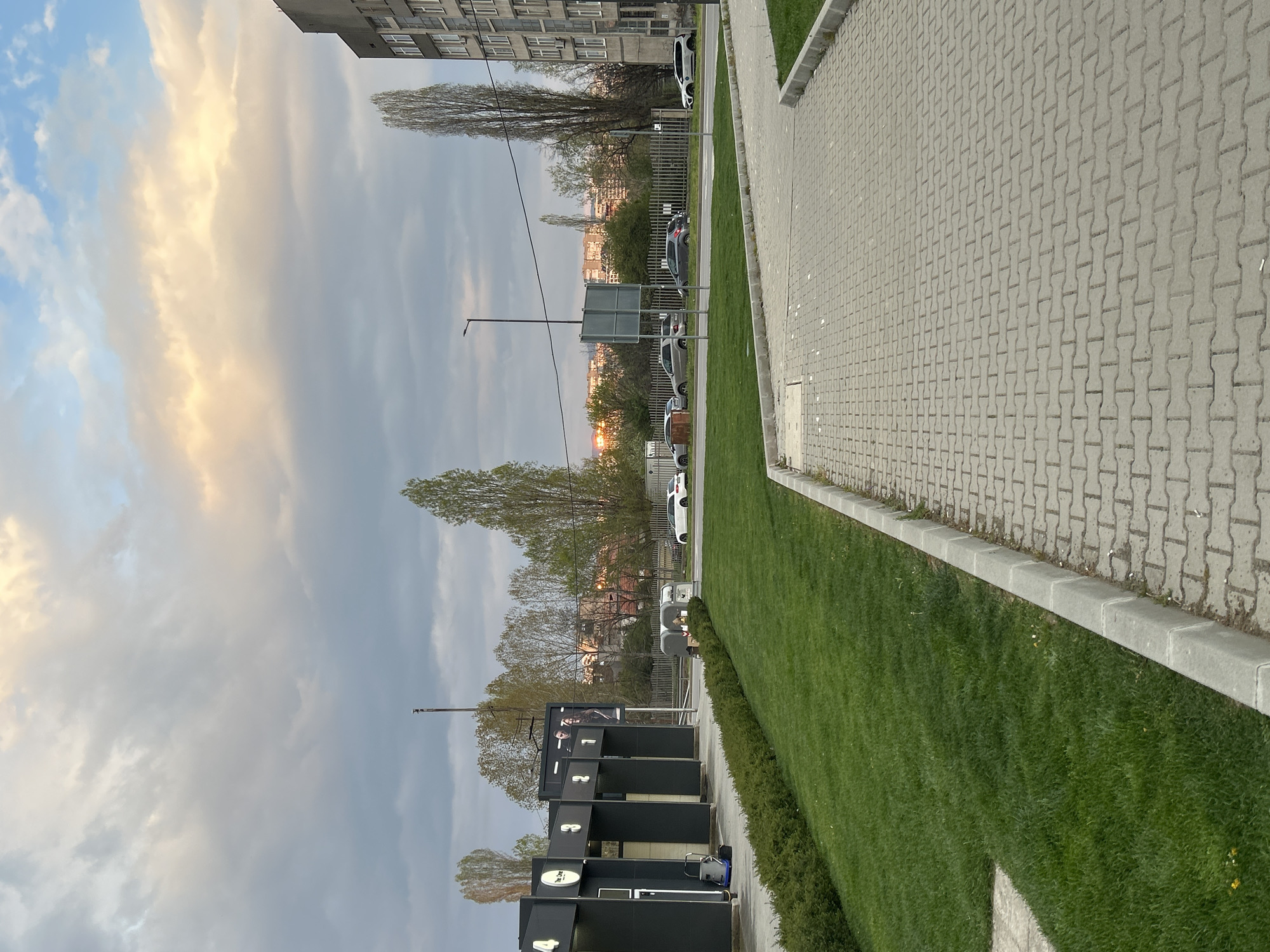 The image displays an urban or suburban street scene, likely in Sofia, Bulgaria, captured during either sunrise or sunset. A patterned concrete block sidewalk forms the foreground, bordered by a strip of manicured green grass. Alongside the grass is a modern-looking, dark-colored structure, possibly a self-service car wash, featuring four numbered bays marked "1," "2," "3," and "4." One bay also has the text "The Car" printed on it. A large billboard on top of this structure shows a person's image. A small blue machine, possibly a vacuum cleaner, is visible near one of the bays.
Beyond this area, a street or parking lot contains several parked cars, including a white SUV and a dark sedan, separated by a metal fence. In the background, a cityscape with buildings and trees stretches towards the horizon, where a bright orange glow indicates the low position of the sun. Tall, slender trees are interspersed throughout the scene. The sky is partly cloudy, with sections of blue and grey clouds catching the warm, golden light of the sun. A lamppost and the back of a street sign are also visible. No specific activity is depicted, and the scene is static.