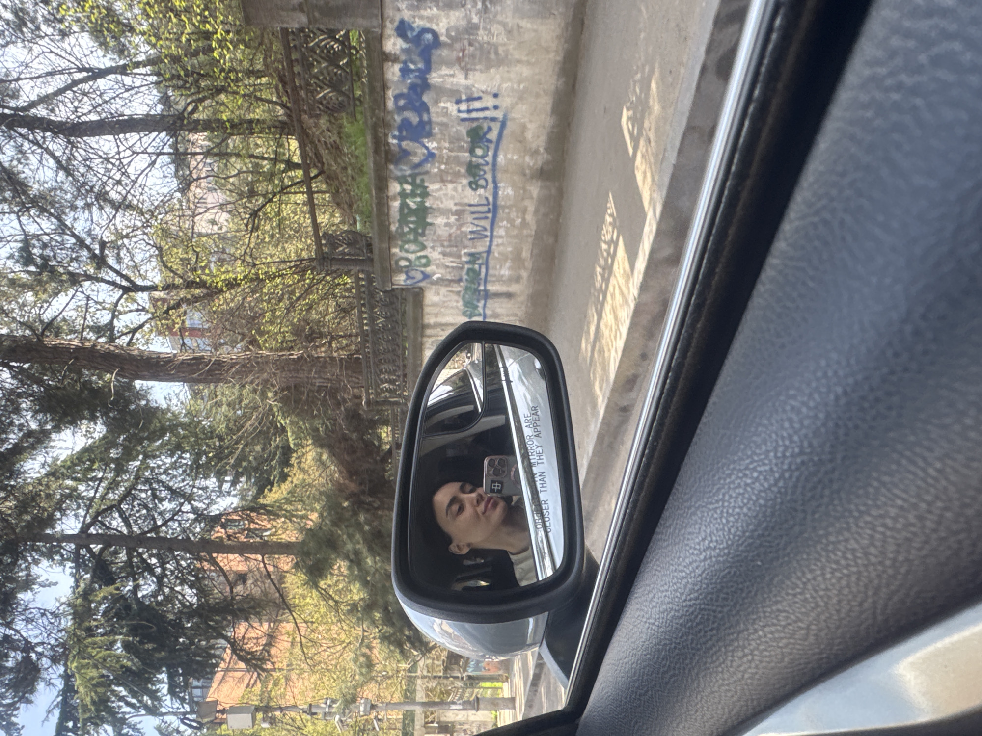 The photograph, taken from inside a car, captures a view reflected in the side mirror and the surrounding environment. In the side mirror, a person is visible, holding a phone to take the picture. The phone case features a character. The mirror also reflects the passing scenery of trees and vegetation. Outside the car, the environment includes trees, a building, and a concrete wall adorned with graffiti. The graffiti includes the words "Women Will Return !!!". The image is likely taken in Tbilisi, Georgia. The car interior is visible, showing a textured, dark-colored surface. The picture seems to be taken during the daytime.