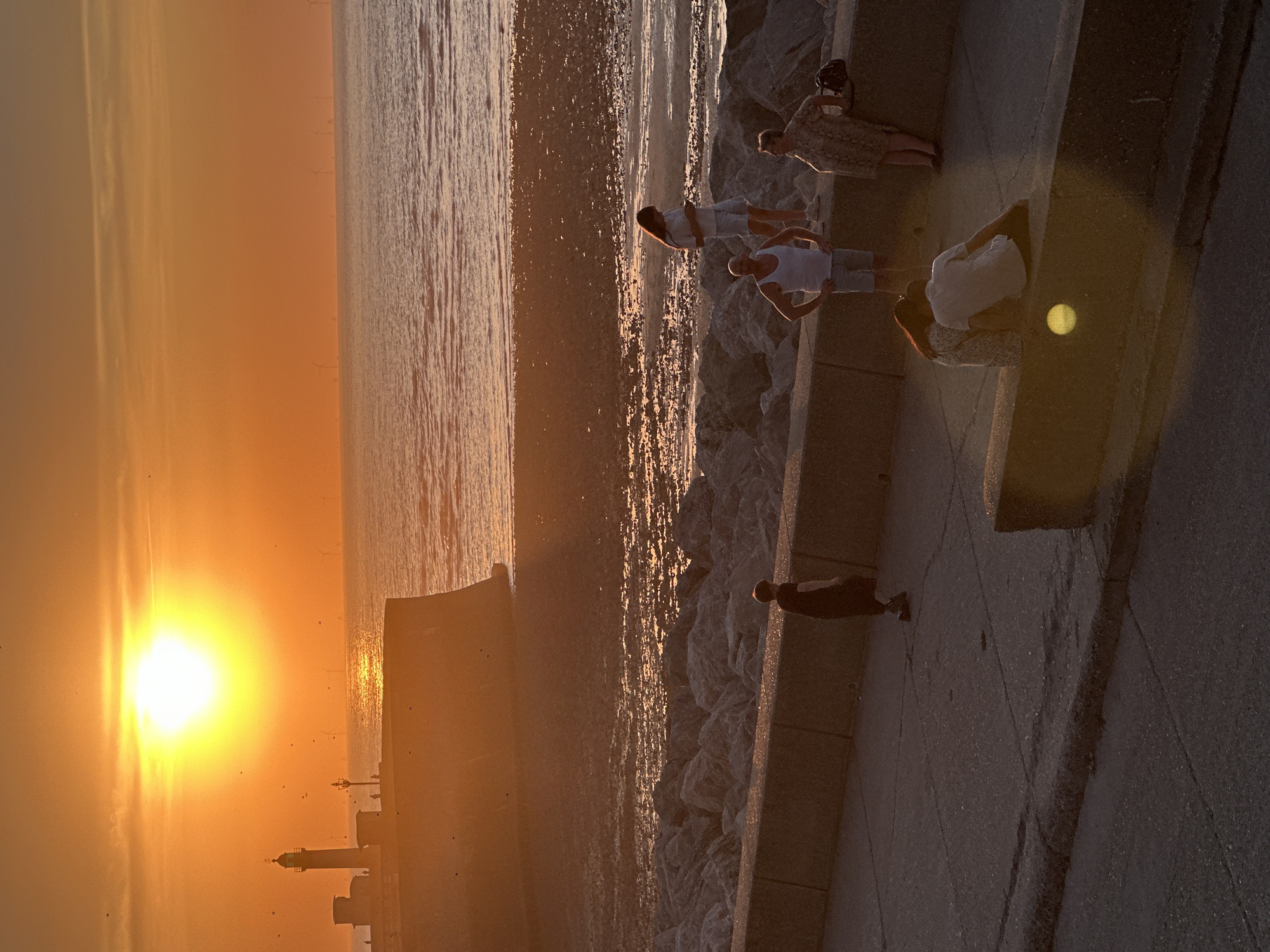 The image depicts a sunset over the sea in Le Tréport, France. A lighthouse stands on a cliff in the background, with a few distant wind turbines visible on the horizon. The foreground shows a concrete promenade with several individuals. On the promenade, a person in a dark dress walks alone. Further along, a couple sits on a low wall, facing away from the camera. Near them, a man in a white tank top and shorts stands with his hands on his hips, looking towards the sea. A woman in a light-colored dress stands nearby, looking at her phone. Another woman in a patterned dress stands beside her. The sea is calm, reflecting the orange and yellow hues of the sunset. The beach area is rocky, with a textured surface of sand and water. The lighting is warm and low, casting long shadows across the scene.