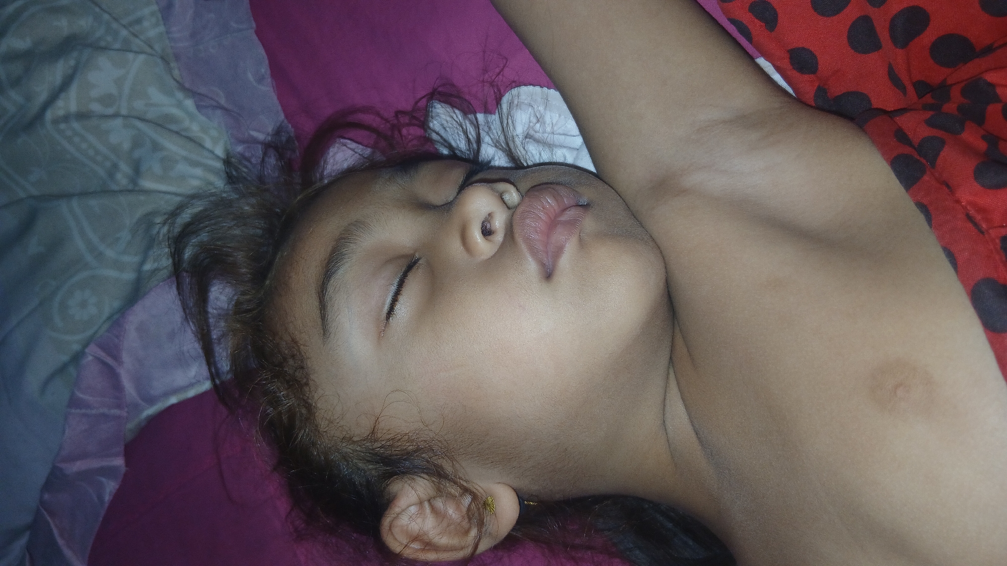 This close-up image features a young child sleeping on a bed. The child is lying on their back, with their head turned to the side. Their eyes are closed, and their lips are slightly parted, with their tongue visible. The child has dark hair, which is spread around their head. They are wearing a small gold earring in their left ear. The background consists of bedding with a patterned grey blanket on the left and a pink blanket underneath. To the right, a red fabric with black polka dots is visible. The lighting suggests it is daytime, likely indoors. The overall mood of the image is peaceful and intimate, capturing a moment of quiet rest. There is no visible text in the image.