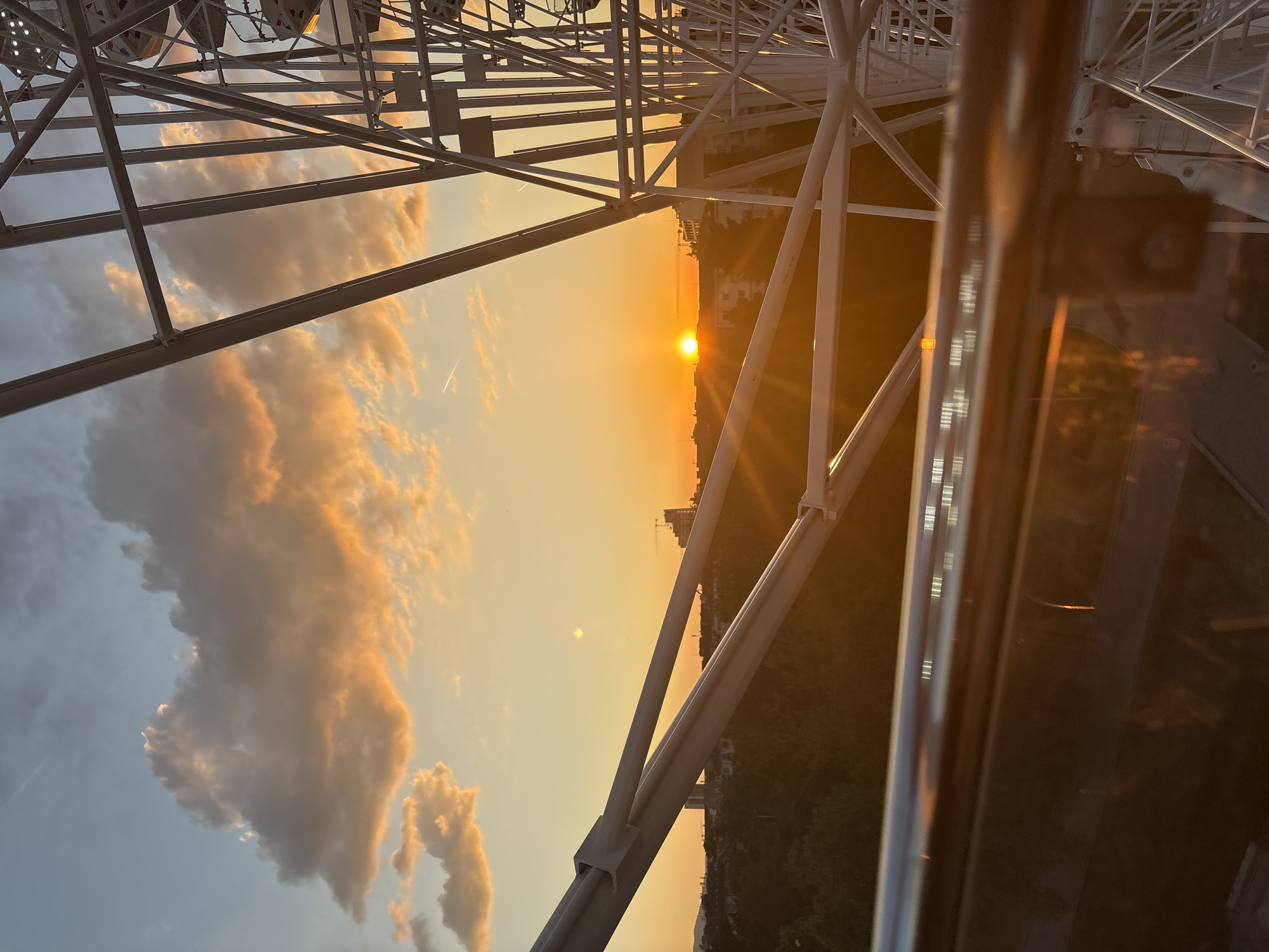 The image depicts a sunset viewed from a Ferris wheel. The sun is setting over the horizon with visible sunbeams. The Ferris wheel structure, made of white metal beams, is prominent in the foreground. Cumulus clouds are visible in the sky, tinged with orange from the setting sun. Below the horizon, there appears to be a city skyline with buildings and green trees. Based on the presence of a Ferris wheel and the described features, the location is likely in Sofia, Bulgaria, specifically the Ferris wheel at Borisova Gradina Park. No people are visible. The image is taken from inside a capsule of the Ferris wheel, which has a reflective surface visible on the right side.