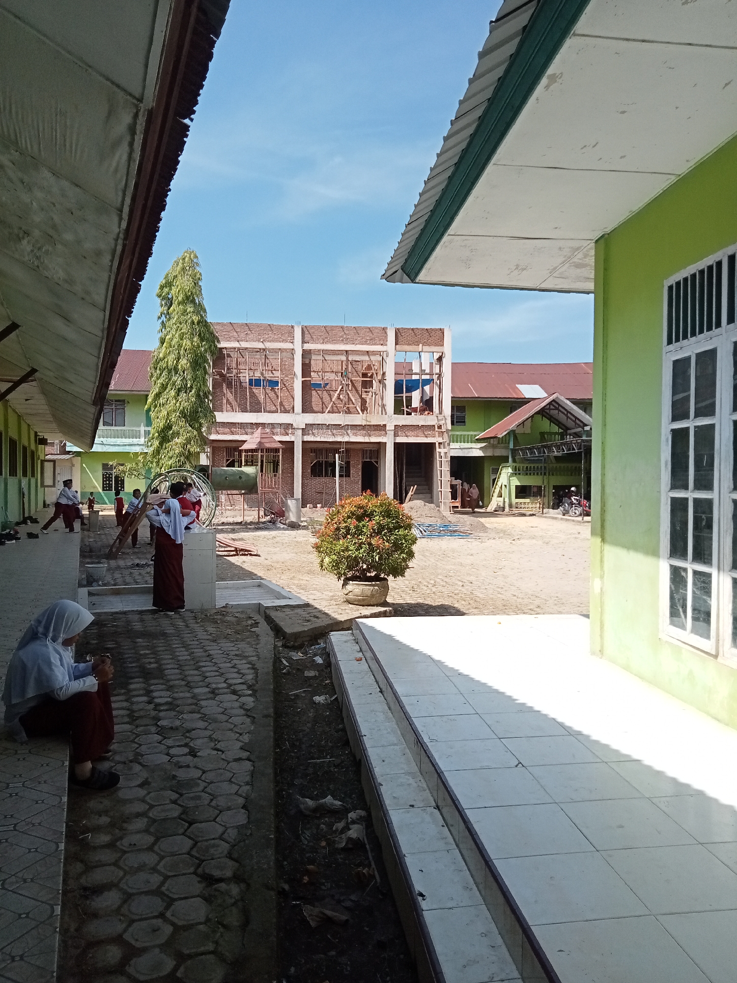 In the city of Kutalhoksukon, Indonesia, a schoolyard under construction is depicted on a clear day. In the foreground, a person wearing a light blue hijab and a maroon skirt is seated on a ledge, looking down at their phone. To their left, another person in similar attire is standing and looking away. The schoolyard features a path paved with hexagonal tiles and a dirt area with building materials, including sand and scaffolding. A partially constructed building with exposed brick and wooden supports stands in the mid-ground, juxtaposed with a mature, tall tree and a bush in a pot. Further back, a green building with a red roof is visible, and a motorcycle is parked near it. The scene is framed by the eaves of two buildings, one on the left with a dark brown roof and another on the right painted bright green with white-framed windows. The image captures a typical day at a school undergoing development.