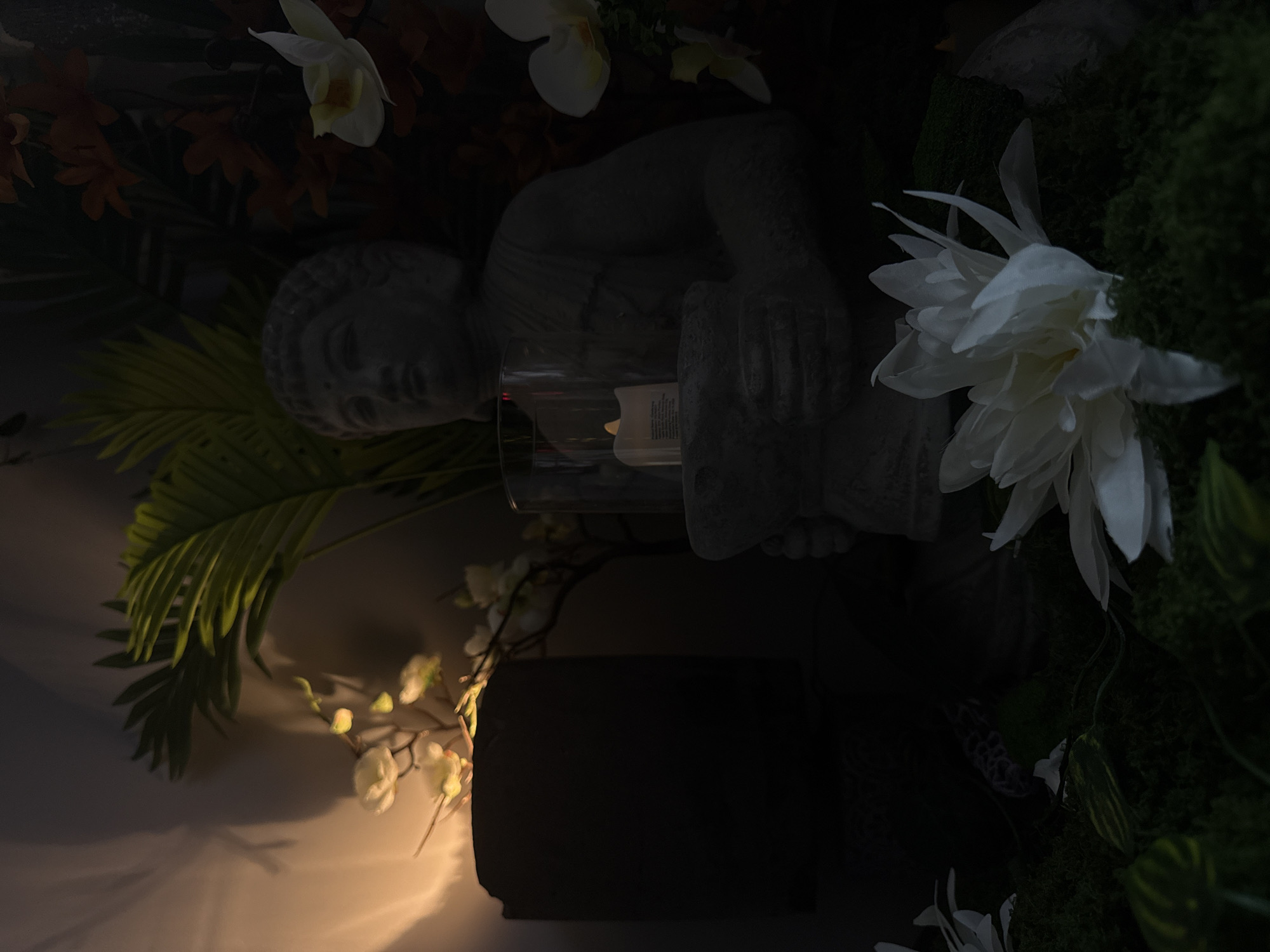 A low-angle, horizontal-oriented photograph shows a stone statue of a reclining figure holding a cylindrical candle holder in a dimly lit setting. The statue is surrounded by artificial plants, including green palm fronds, white and yellow orchids, and green moss. The light source, a candle within the holder, casts a warm glow on the objects below, illuminating a dark pedestal. The setting is indeterminate. No people or interactions are visible. There are no specific indicators to place the scene in Petarch, Bulgaria.