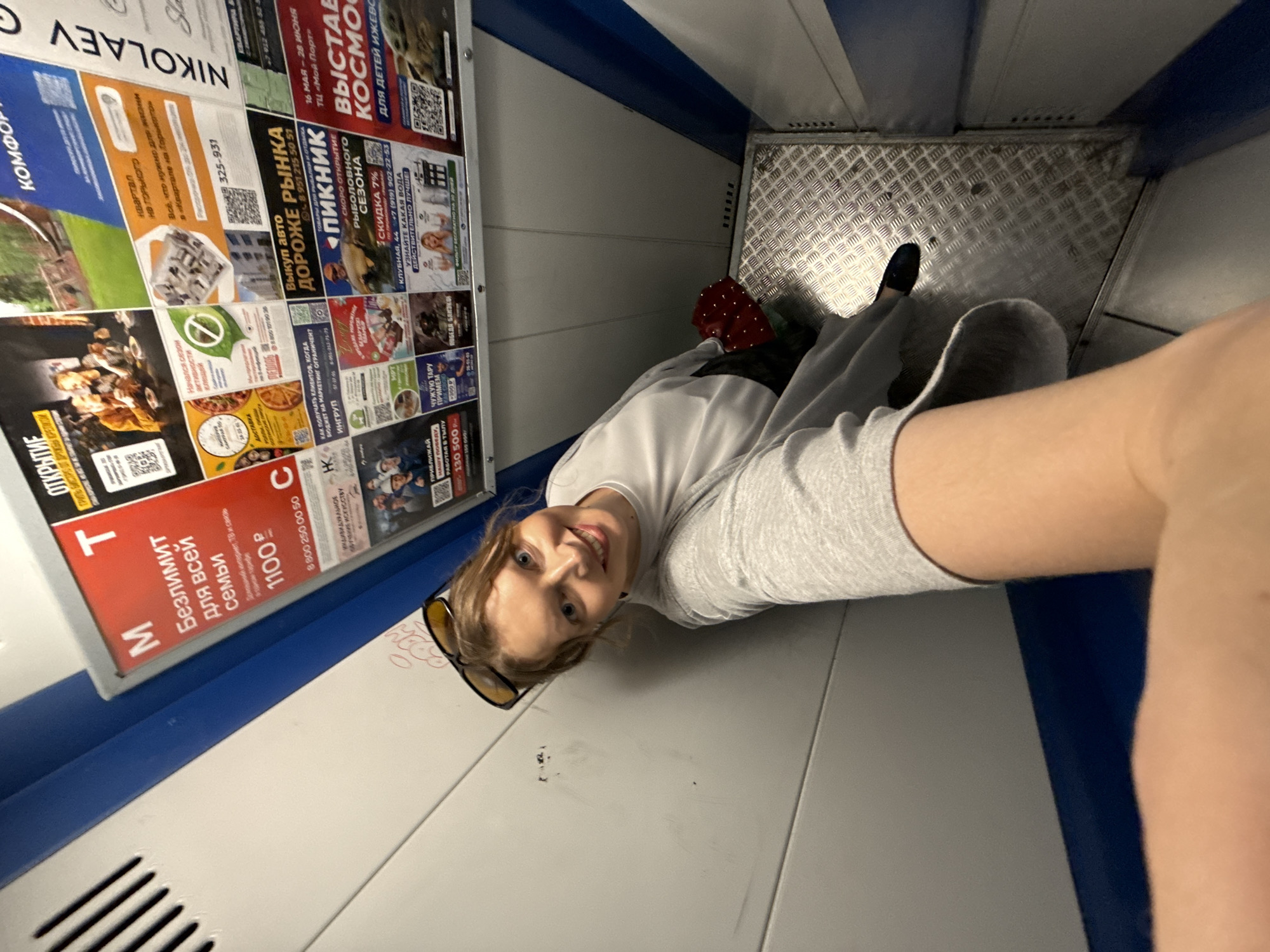A woman is sitting in an elevator in Izhevsk, Russia. She is wearing a white t-shirt and grey sweater and sunglasses. The elevator has advertisements on one wall, including ads for real estate, auto sales, and a fishing season opening. One ad references "Nikolaev." The floor of the elevator is metal with a diamond pattern.