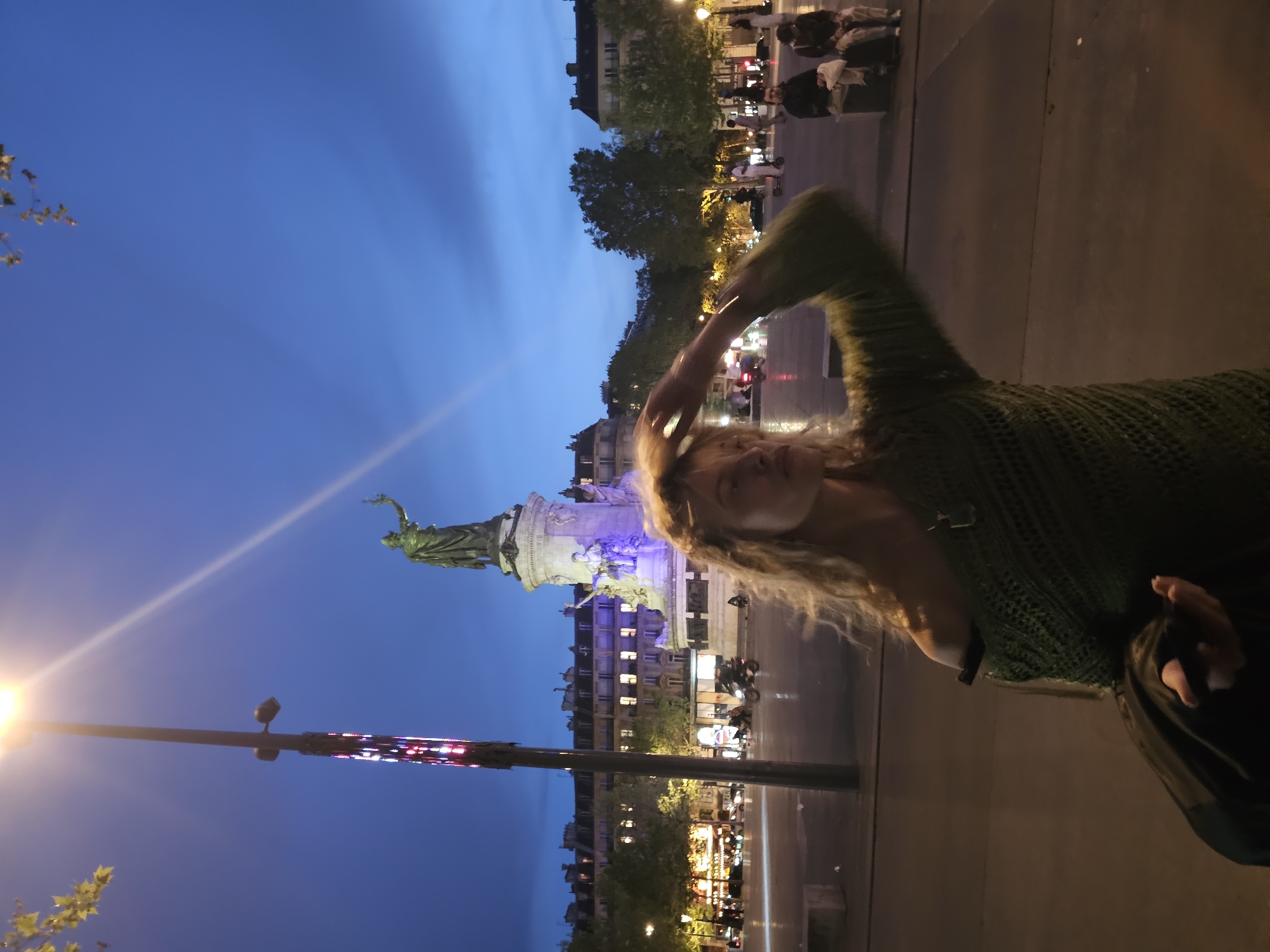 At twilight, the Place de la République in Paris, France, serves as the backdrop for a posed photo of a Caucasian woman with long, blonde hair. She wears a dark green knit top and holds her hand near her head. In the background, the statue of Marianne stands illuminated. Traffic moves along the periphery of the square, and buildings flank the area. A streetlight flares near a decorative pole. Pedestrians are visible in the upper right corner of the frame. The pavement is visible in the foreground.