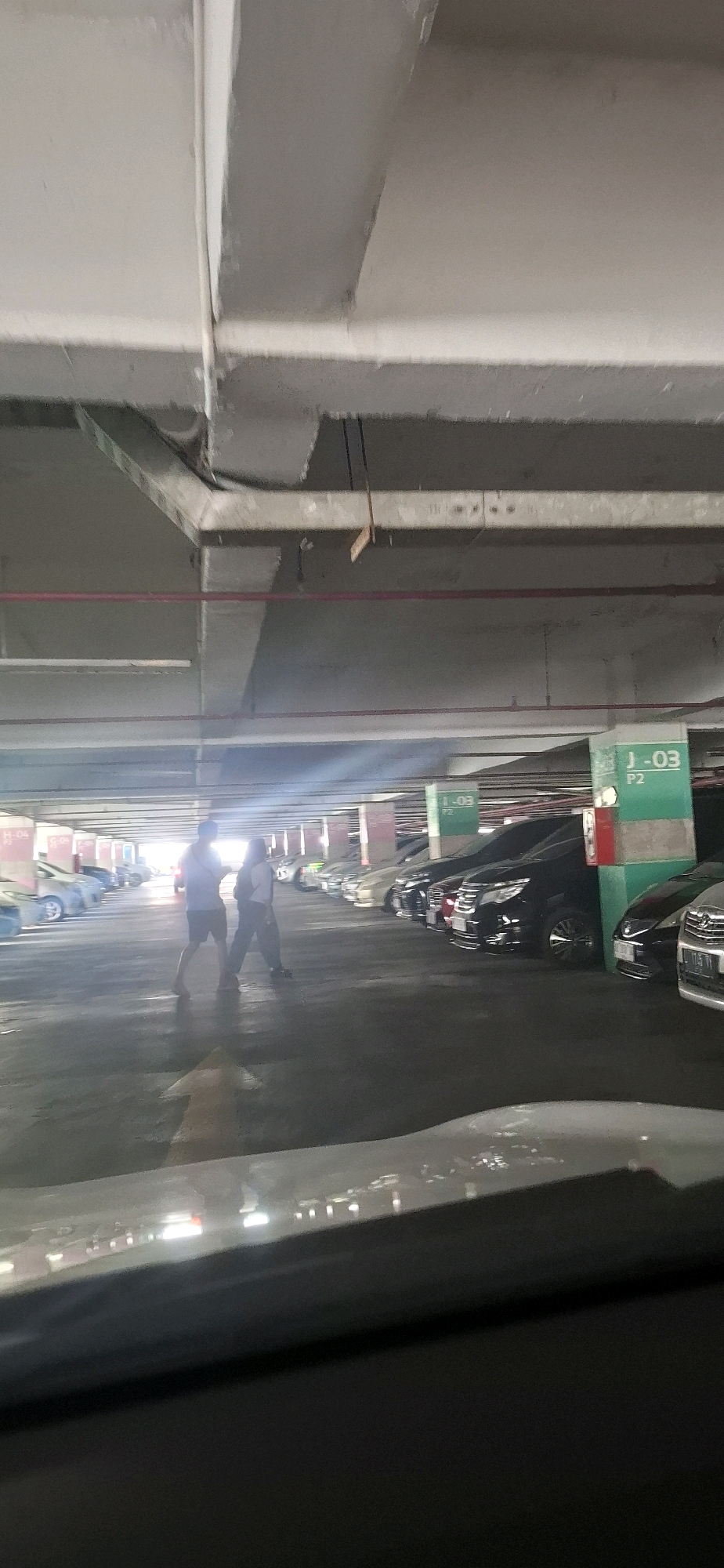 This is a photograph taken inside a multi-level parking garage, likely in the city of Surakarta, Indonesia, given the context. The image is characterized by a wide-angle perspective, emphasizing the concrete structure of the garage. Visible in the foreground is the interior of a vehicle, with a blurred dashboard and windshield.
The scene depicts several parked cars, primarily SUVs and sedans, arranged in rows. The lighting suggests artificial illumination from the garage's fixtures, with some natural light filtering in from the far end, creating a gradient of brightness. There are concrete pillars, some painted with section markers such as "J-03 P2" and "H-04 G-05". A red pipe runs horizontally across the ceiling structure.
In the mid-ground, two individuals are walking across the parking aisle. One appears to be a male wearing a light-colored t-shirt and dark shorts, while the other is a female in a dark top and light-colored pants. They are walking towards the right side of the frame, away from the viewer. The overall atmosphere is typical of an indoor parking facility.