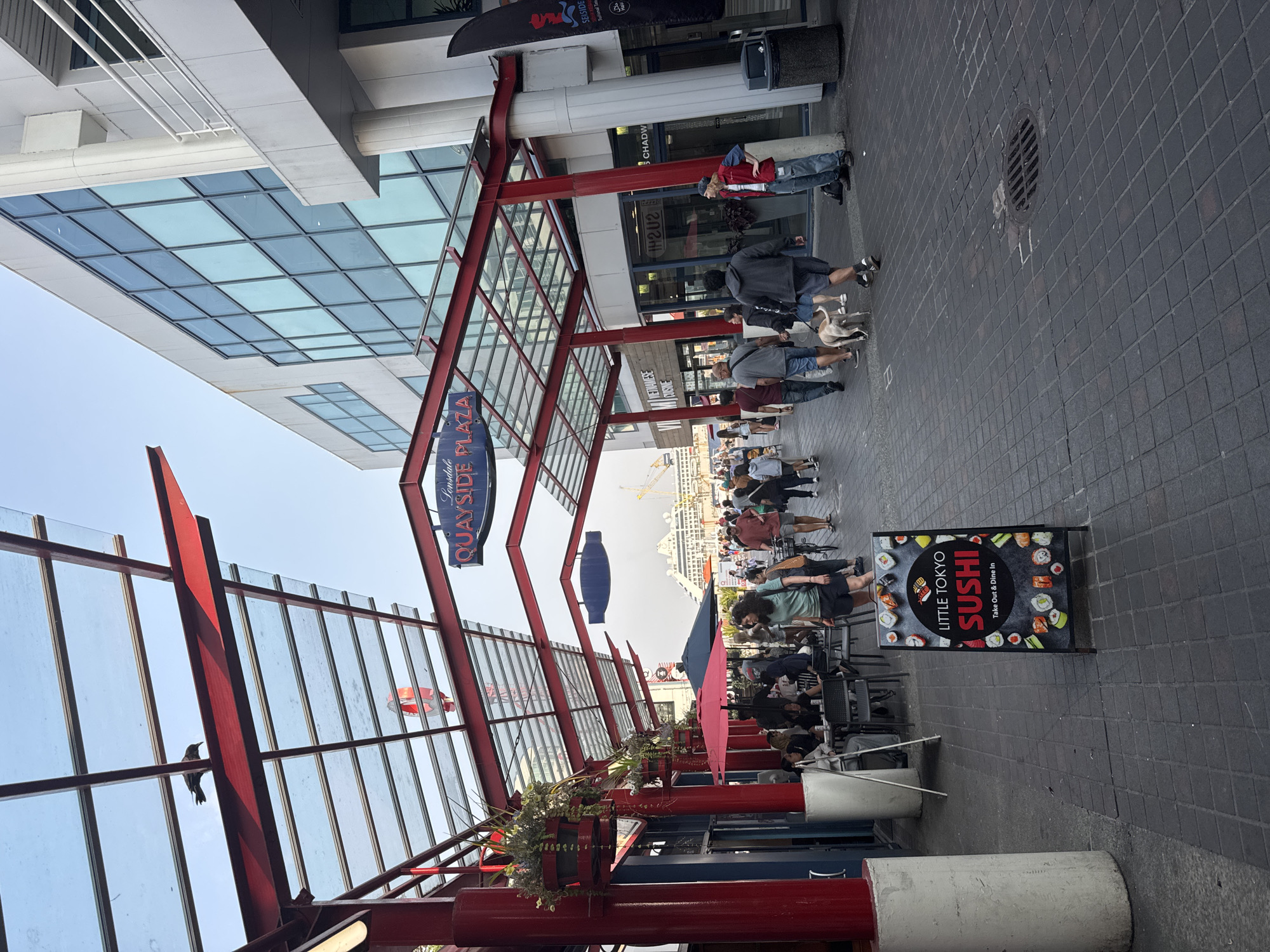 The image shows a street scene at Lonsdale Quay Plaza in North Vancouver, Canada. The foreground features a red metal and glass canopy structure with white cylindrical pillars. Several people are walking along the paved walkway, some with dogs. To the left, there are outdoor seating areas for a restaurant, indicated by tables and chairs. A sign for "Little Tokyo Sushi" is prominently displayed on the right side of the walkway. In the background, a large cruise ship is docked, and construction cranes are visible, suggesting ongoing development or activity at the waterfront. The architecture of the buildings lining the plaza is modern, with large glass windows. The sky is clear and bright.
