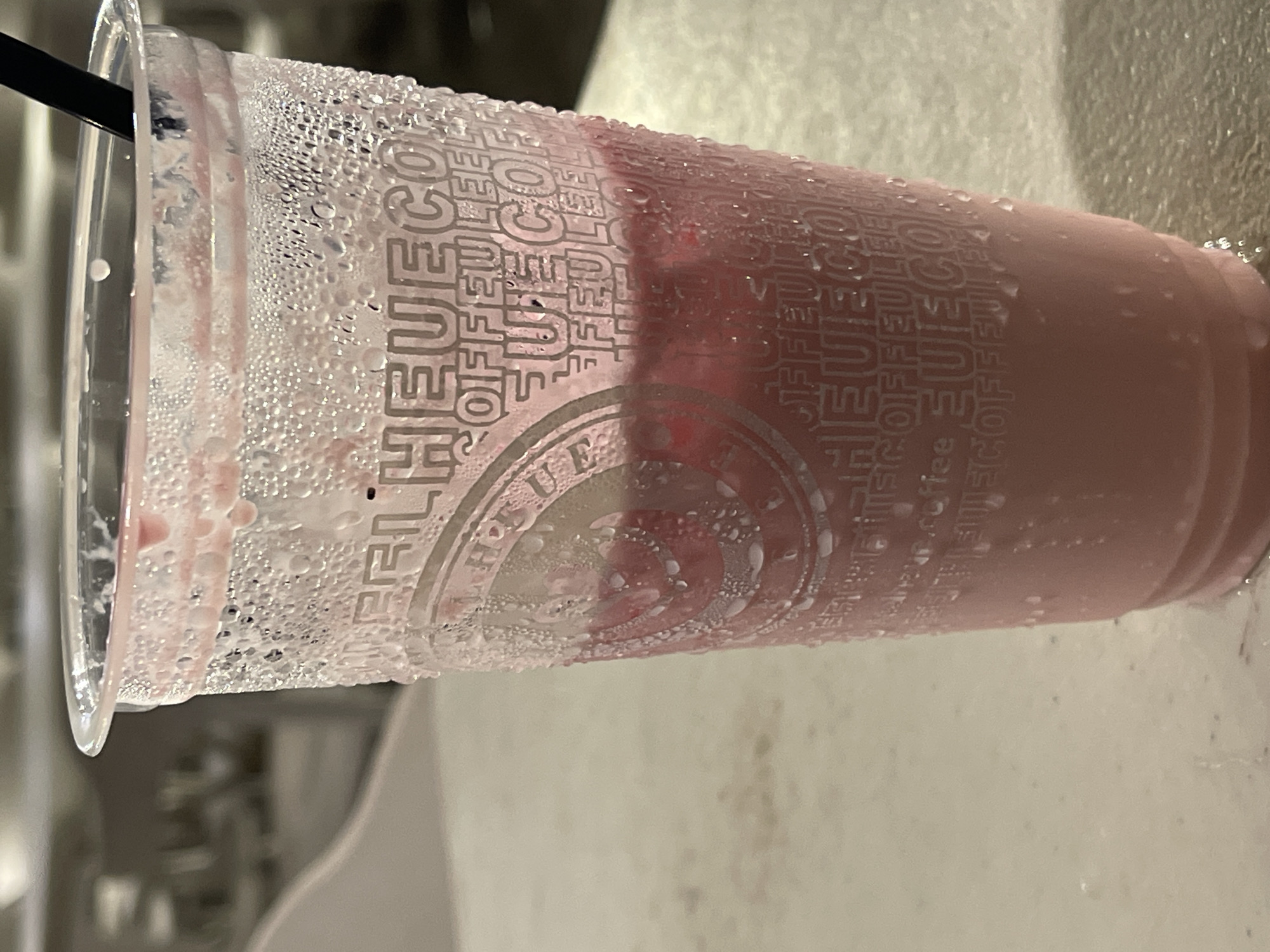 A close-up shot shows a clear plastic cup filled with a pink beverage, likely a strawberry smoothie or milkshake. The outside of the cup is covered in condensation, with droplets of water clinging to its surface. The cup has a repeating pattern of text and logos, partially obscured by the condensation. A black straw is visible inserted into the drink. The cup rests on a light-colored, possibly textured surface, and the background is blurred, suggesting an indoor or dimly lit outdoor setting. The overall impression is of a refreshing, cold drink.