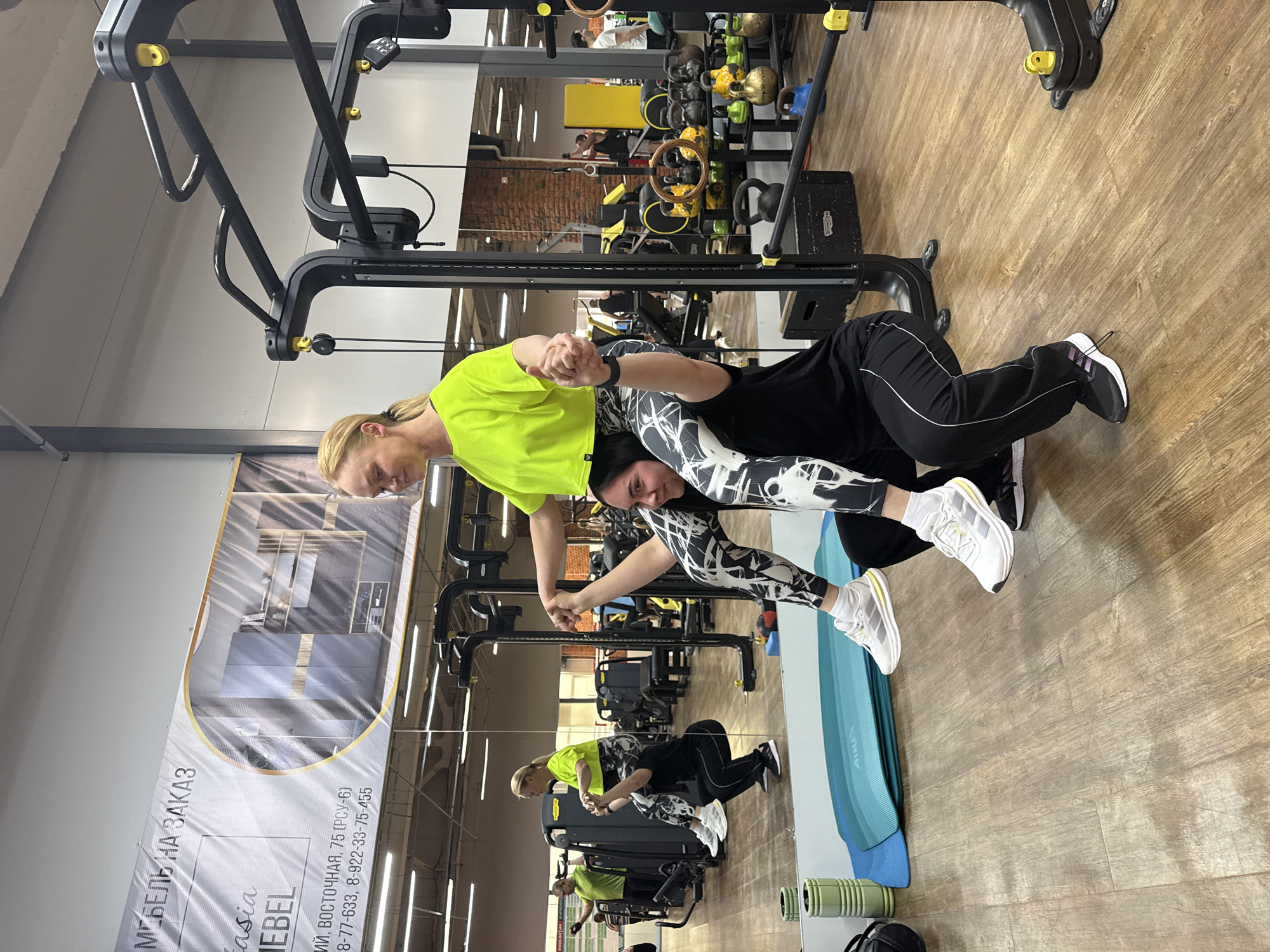 This image, rotated 90 degrees clockwise, captures two women engaging in a partner exercise or stretch within a well-lit gym setting. The woman on the left, wearing a vibrant yellow-green t-shirt and dark athletic pants, holds hands with the woman on the right. The second woman is dressed in a black top and distinctive black-and-white patterned leggings, positioned in a crouching or bent-over stance, seemingly supported by her partner. Both are wearing athletic shoes.
The gym features a light wooden floor and various workout equipment in the background, including a black functional trainer machine, a rack filled with kettlebells of different colors, and exercise mats on the floor. A large mirror on the right reflects the scene, showing the women and other gym elements, including a person exercising further in the background. The woman in patterned leggings looks towards the camera with a slight smile, while her partner is looking downwards, focused on the activity.
On the left side, a partially visible banner or advertisement in Russian displays text: "МЕБЕЛЬ НА ЗАКАЗ" (Custom Furniture), "asia EBEL," followed by an address fragment "ИЙ, ВОСТОЧНАЯ, 75 (РСУ-6)," and phone numbers "8-77-633" and "8-922-33-75-455." This detail, combined with the location context of Chaikovsky, Russia, suggests the gym may be situated at Vostochnaya 75, or a nearby location, potentially sharing premises or advertising with a furniture business. The indoor lighting indicates it is likely daytime.