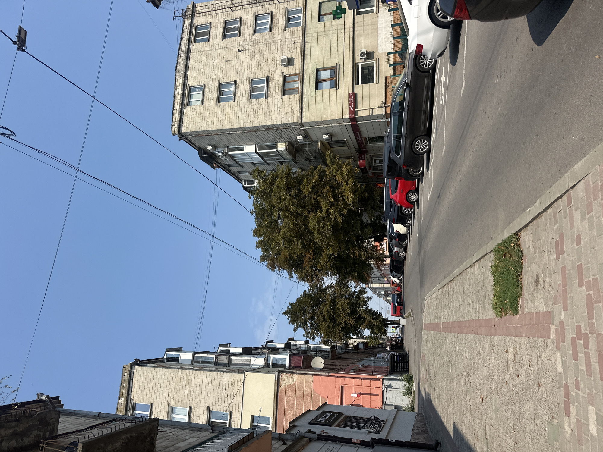 The image depicts a street scene in Ternopil, Ukraine, on a clear day. The perspective is from a sidewalk looking down a street lined with multi-story buildings and parked cars. On the left, a faded, pink-colored building with multiple windows and balconies is visible, adjacent to a lighter-colored brick building with a similar facade. The sidewalk is paved with stone blocks, with a decorative red brick inlay. On the right side of the street, several cars are parked, including a dark-colored station wagon, a red hatchback, and a white sedan. A taller, light-colored brick building with numerous windows and balconies dominates the right side of the street. Tall, leafy trees are situated between the buildings, casting shadows. Overhead, a network of power lines crisscrosses the sky. The street itself is paved with asphalt and marked with white directional arrows. A green cross symbol is visible on the right side of the street, likely indicating a pharmacy. The overall impression is one of an urban environment with residential and commercial buildings, common in Eastern European cities.
