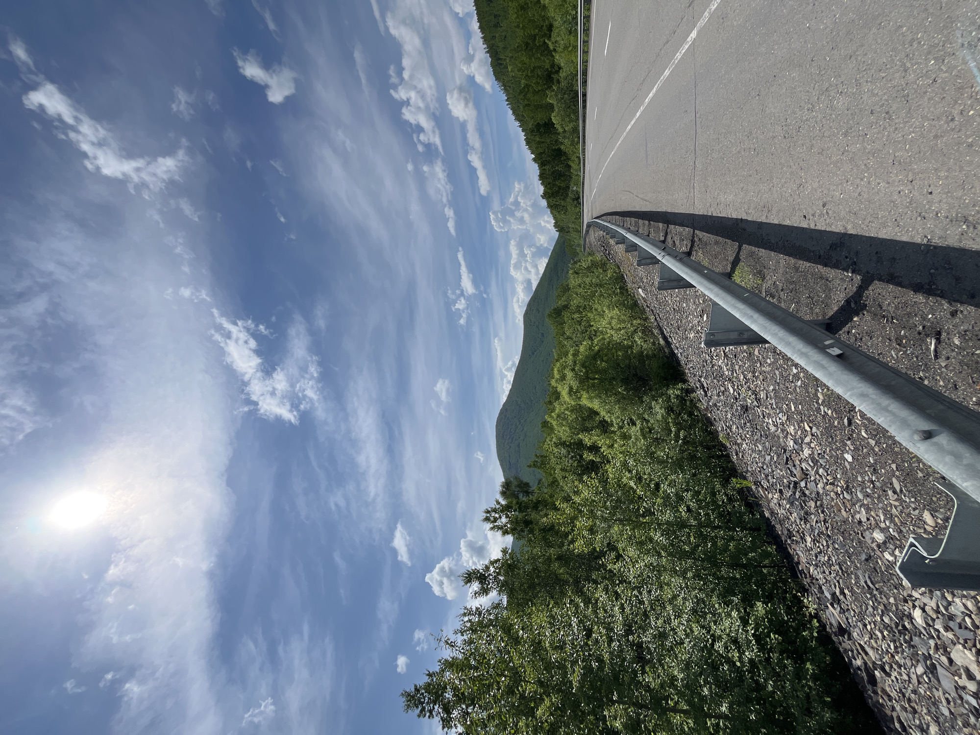 The image depicts a scenic view of a highway in a mountainous, forested region. A paved road curves gently to the right, bordered by a metal guardrail and a rocky embankment covered in small stones. Lush green trees and dense foliage line the left side of the road, extending up the slopes of rolling, green hills. The sky above is a vibrant blue, dotted with wispy white clouds. The sun is high and bright, casting a strong light and shadows. The overall impression is one of a serene and natural landscape, possibly in a rural or undeveloped area of Russia.