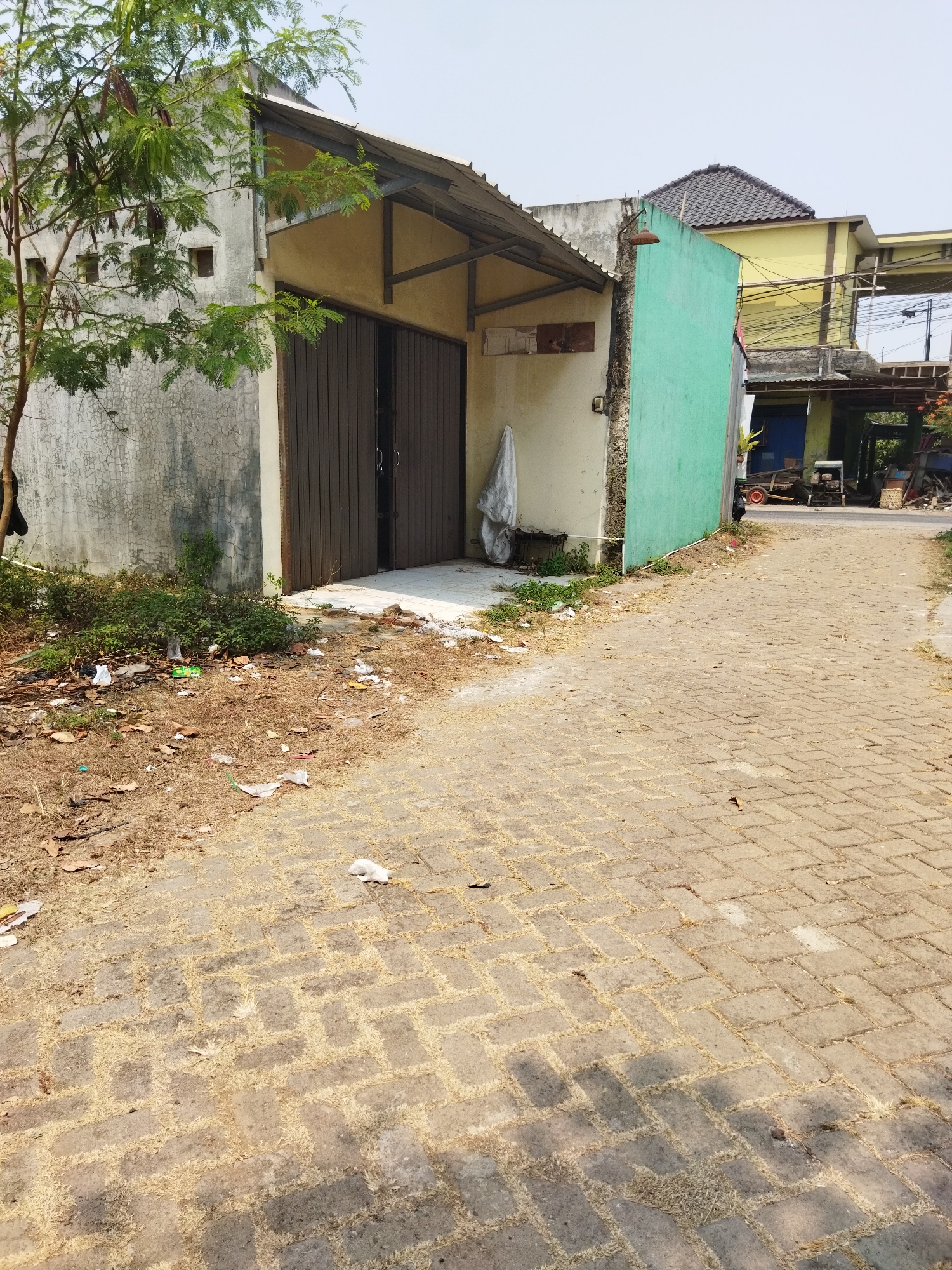 This is an outdoor, eye-level, full shot image taken on a sunny day in Suka Sari, Indonesia. The scene depicts a paved walkway leading past a building with a faded grey concrete exterior and a section of bright green painted wall. The building has a corrugated metal roof supported by wooden beams. In front of the building, there is a set of large, dark brown wooden doors. To the left of the building, a small tree with green leaves and pods is growing. The ground next to the walkway is covered with dry grass, fallen leaves, and scattered litter, including white plastic bags and other debris. Further down the walkway, other buildings are visible, including a yellow building with a dark grey tiled roof and a structure with a blue door. In the background, there are some vehicles, including what appears to be a small trailer with red wheels and a larger piece of machinery. The sky is mostly clear with a few faint clouds. The overall impression is one of a somewhat neglected urban or semi-urban environment.