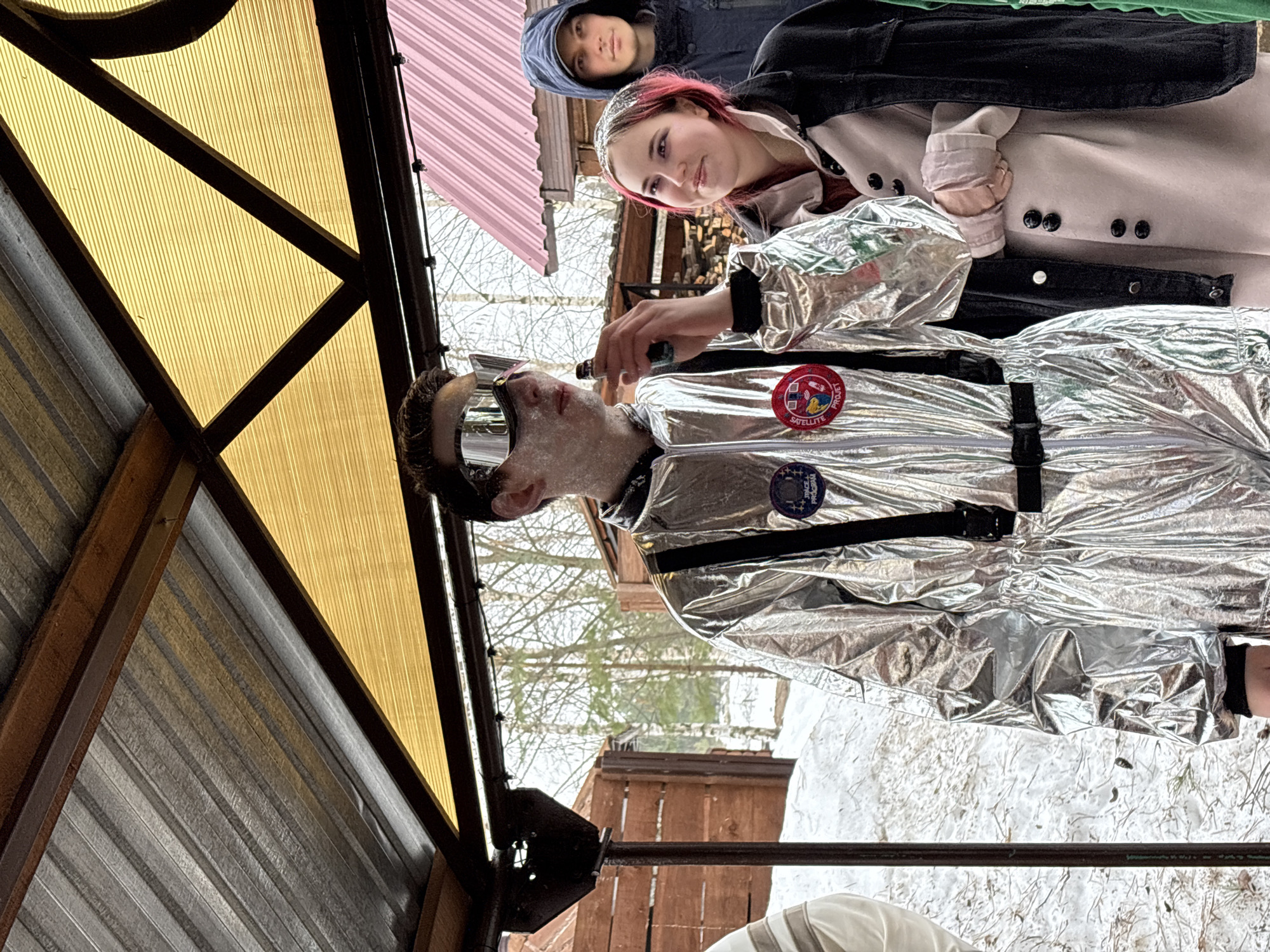 Outside in winter, three people are gathered near a structure with a yellow translucent roof and metal corrugated siding, possibly in or near Chaikovsky, Russia. The central figure wears a silver jumpsuit with patches and goggles, applying something from a small bottle to their face. Behind them, a person with pink hair in a light-colored coat stands alongside another person in a blue hooded jacket. The ground is covered in snow.